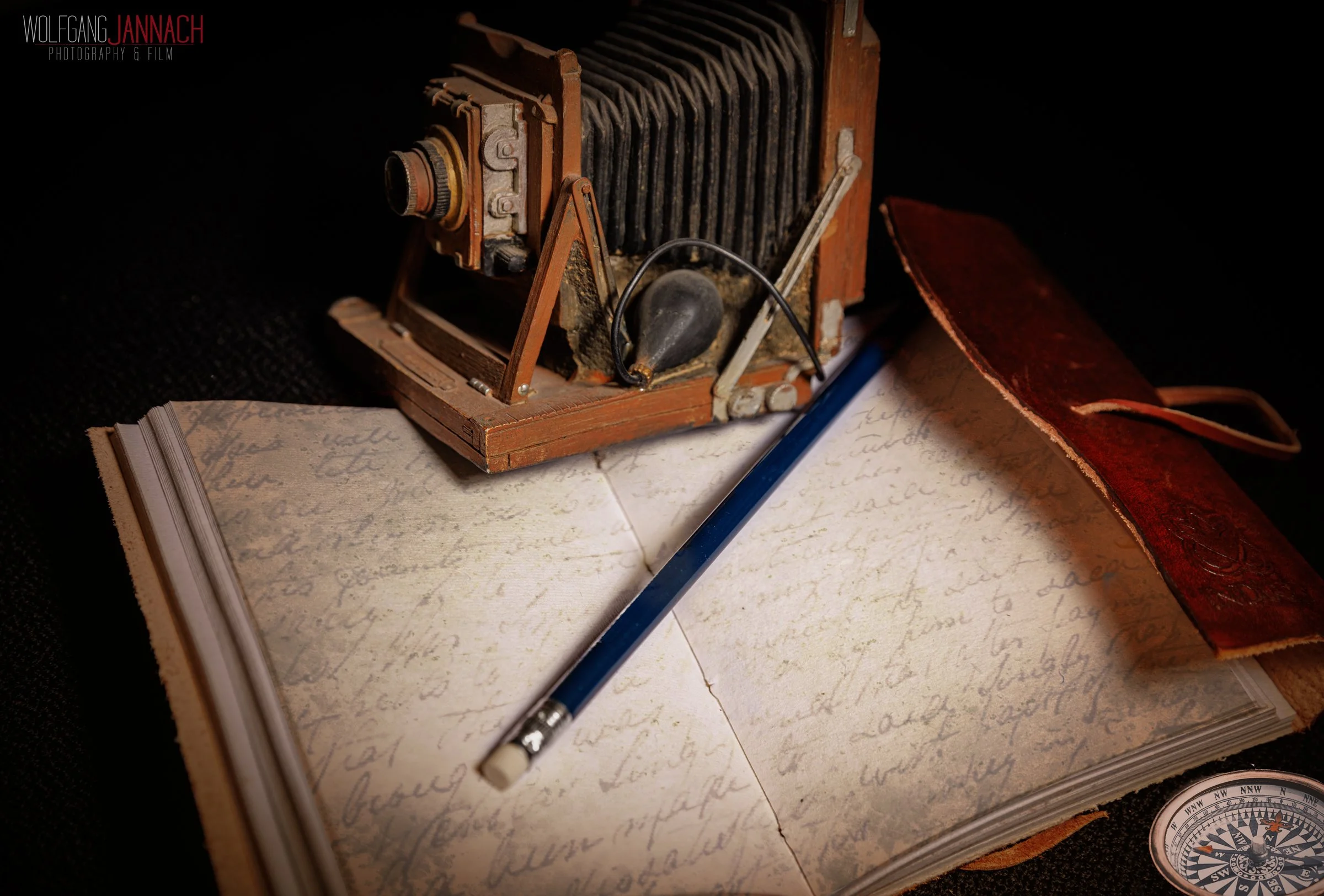 Ein altes, vintage Fotoapparat, ein geöffnetes Notizbuch mit handgeschriebenem Text, ein blauer Bleistift, ein rotes Filzhülle, ein Kompass auf schwarzem Hintergrund.