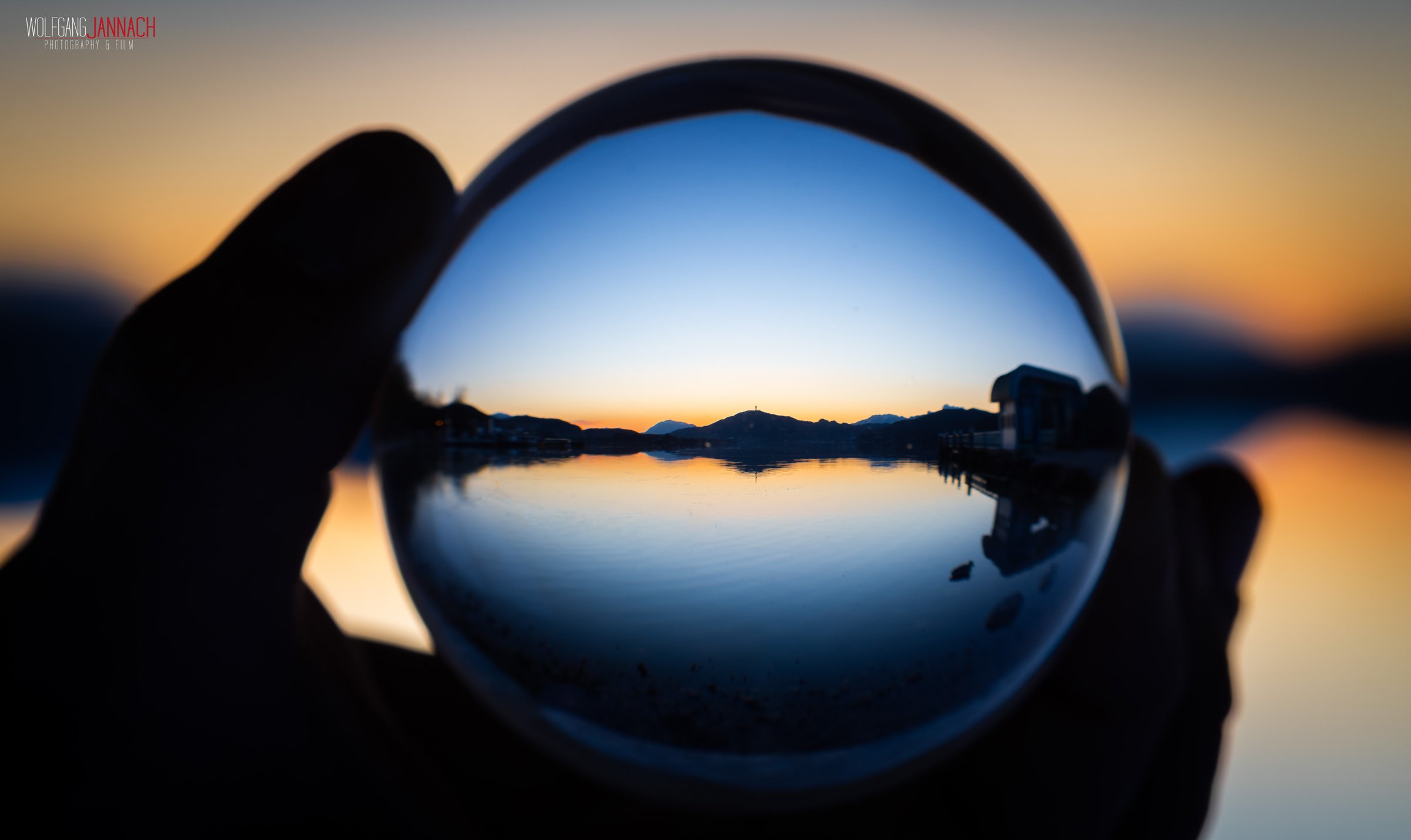 Sicht auf einen ruhigen See bei Sonnenuntergang, gesehen durch eine Brille, mit Bergen im Hintergrund.