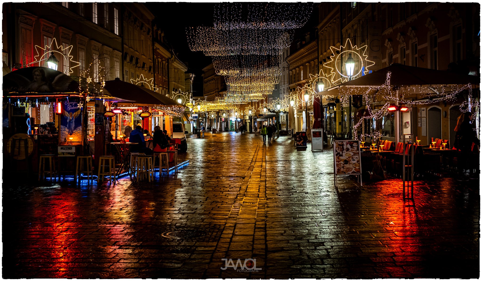Eine leere, regennasse, beleuchtete Fußgängerzone mit festlicher Weihnachtsbeleuchtung und Marktständen auf beiden Seiten bei Nacht.