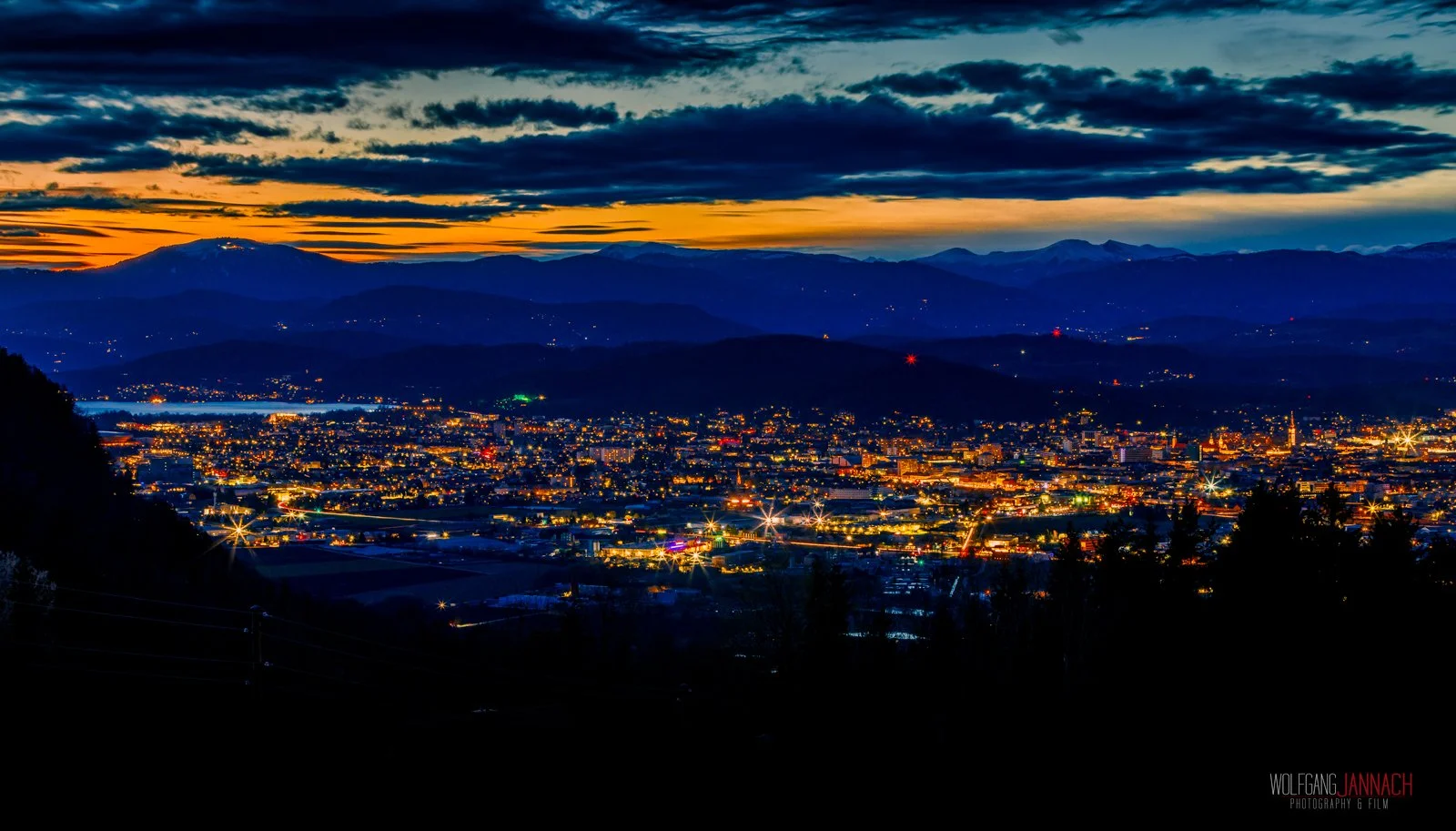 Eine nächtliche Stadtansicht mit leuchtenden Lichtern, umgeben von Bergen unter einem bewölkten Himmel bei Sonnenuntergang oder Dämmerung.