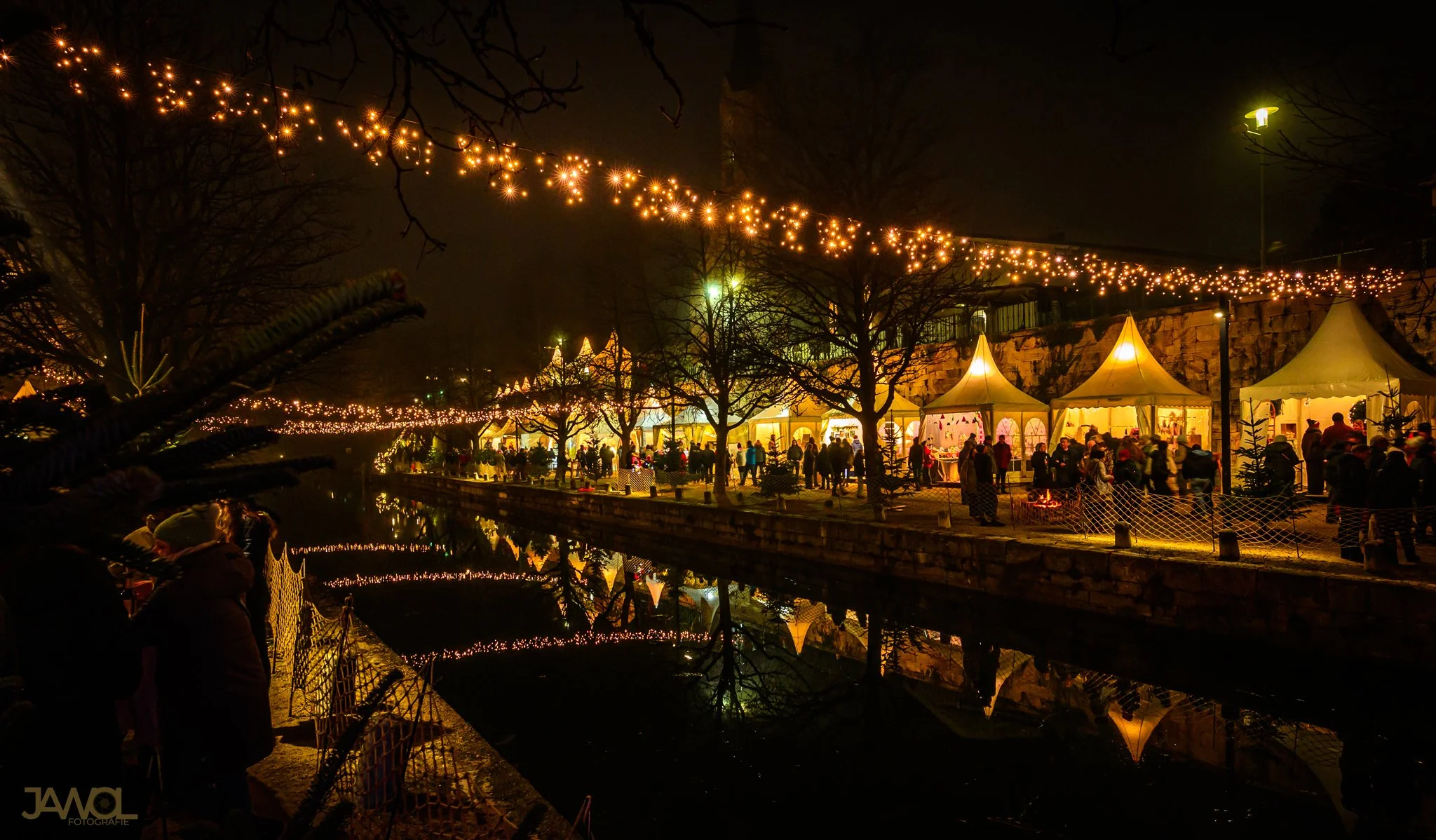 Weihnachtsmarkt am Fluss bei Nacht mit leuchtenden Lichterketten, Zelten und Besucher