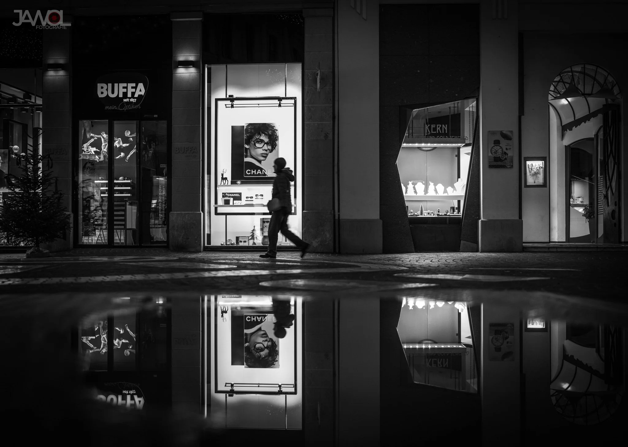 Straßenszene bei Nacht, ein Mensch läuft vor einem Schaufenster mit Werbung für Brillen, im Hintergrund leuchtet eine Boutique. Es gibt eine Wasserpfütze, die die Szene reflektiert.