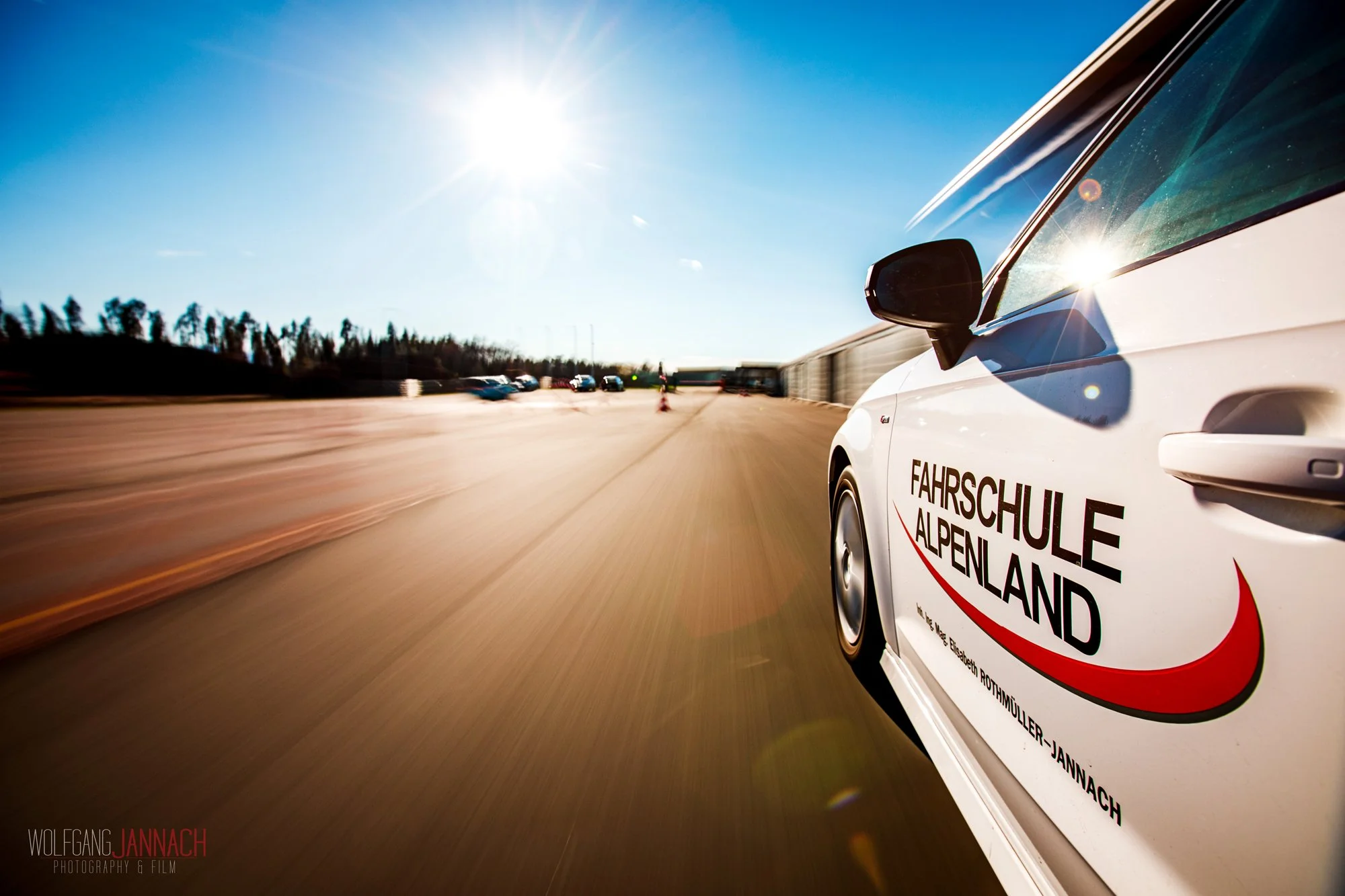 Weiße Fahrschulauto mit dem Logo 'Fahrschule Alpenland' fährt auf einer offenen Strecke bei Sonnenschein, im Hintergrund sind weitere Fahrzeuge und Bäume sichtbar.