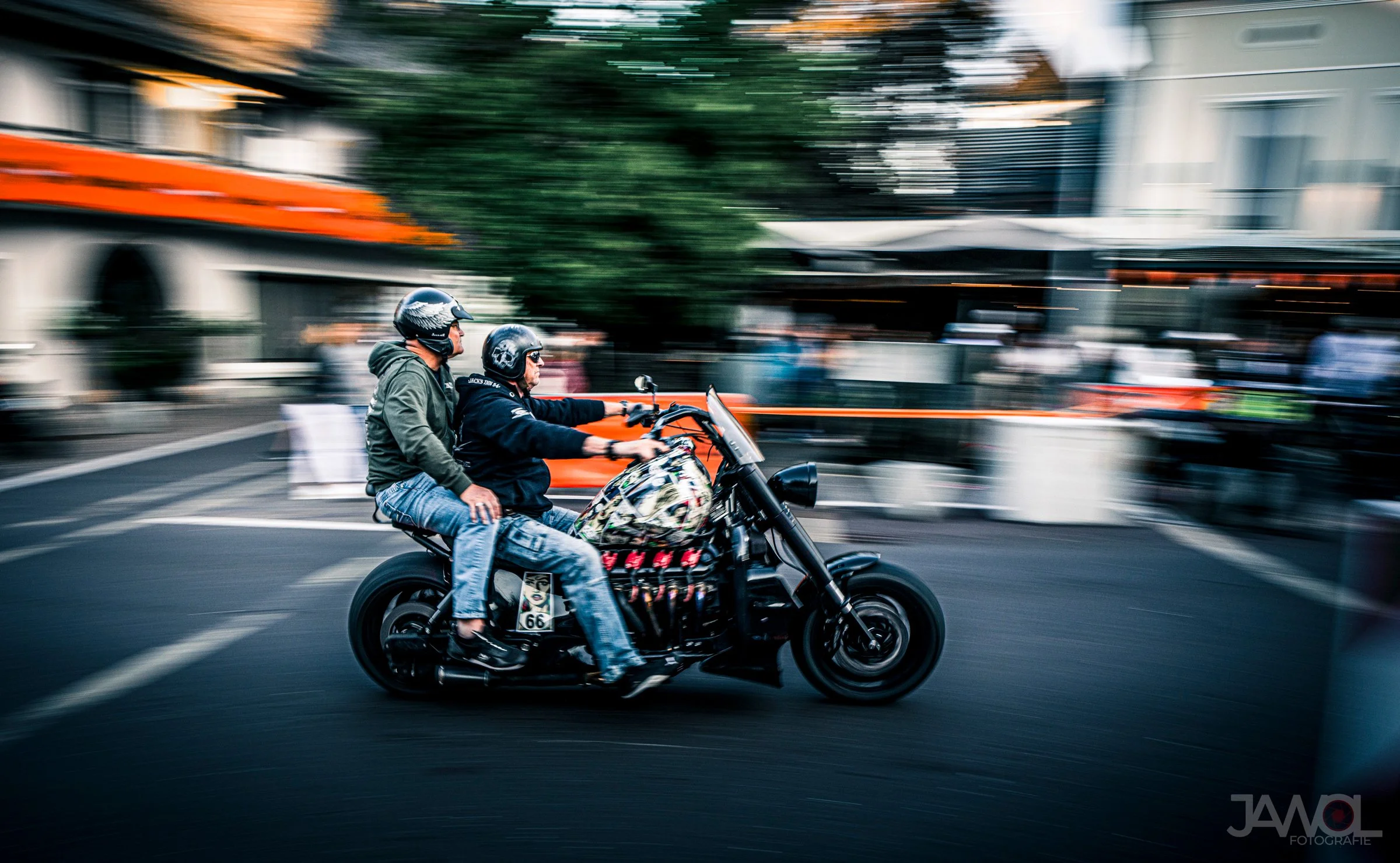 Zwei Männer auf einem schwarzen Motorrad fahren durch eine geschäftige Straße, während die Umgebung in Bewegung unscharf ist.