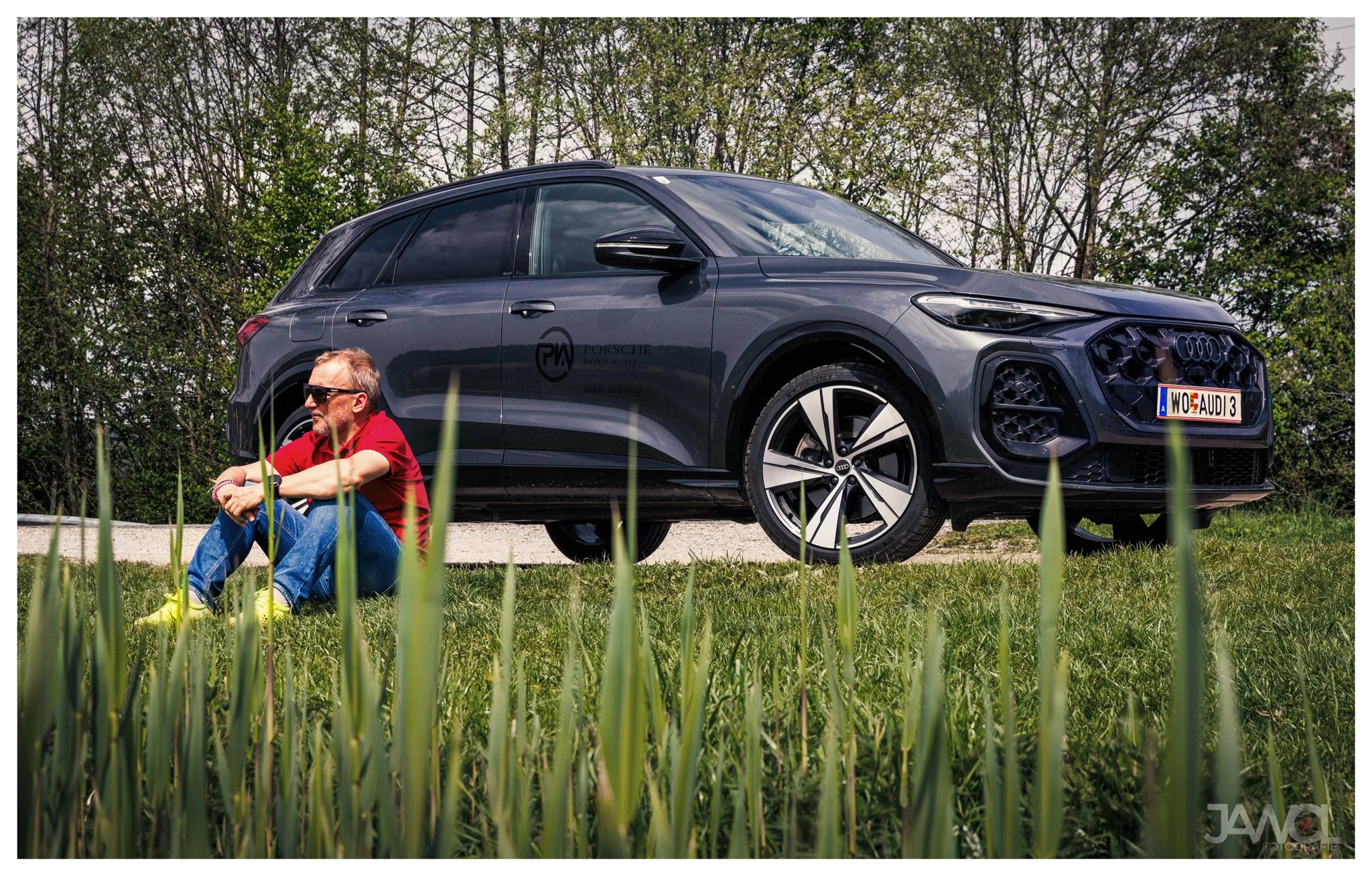 Ein Mann sitzt entspannt auf dem Gras vor einem schwarzen Audi-Auto, im Hintergrund Bäume.
