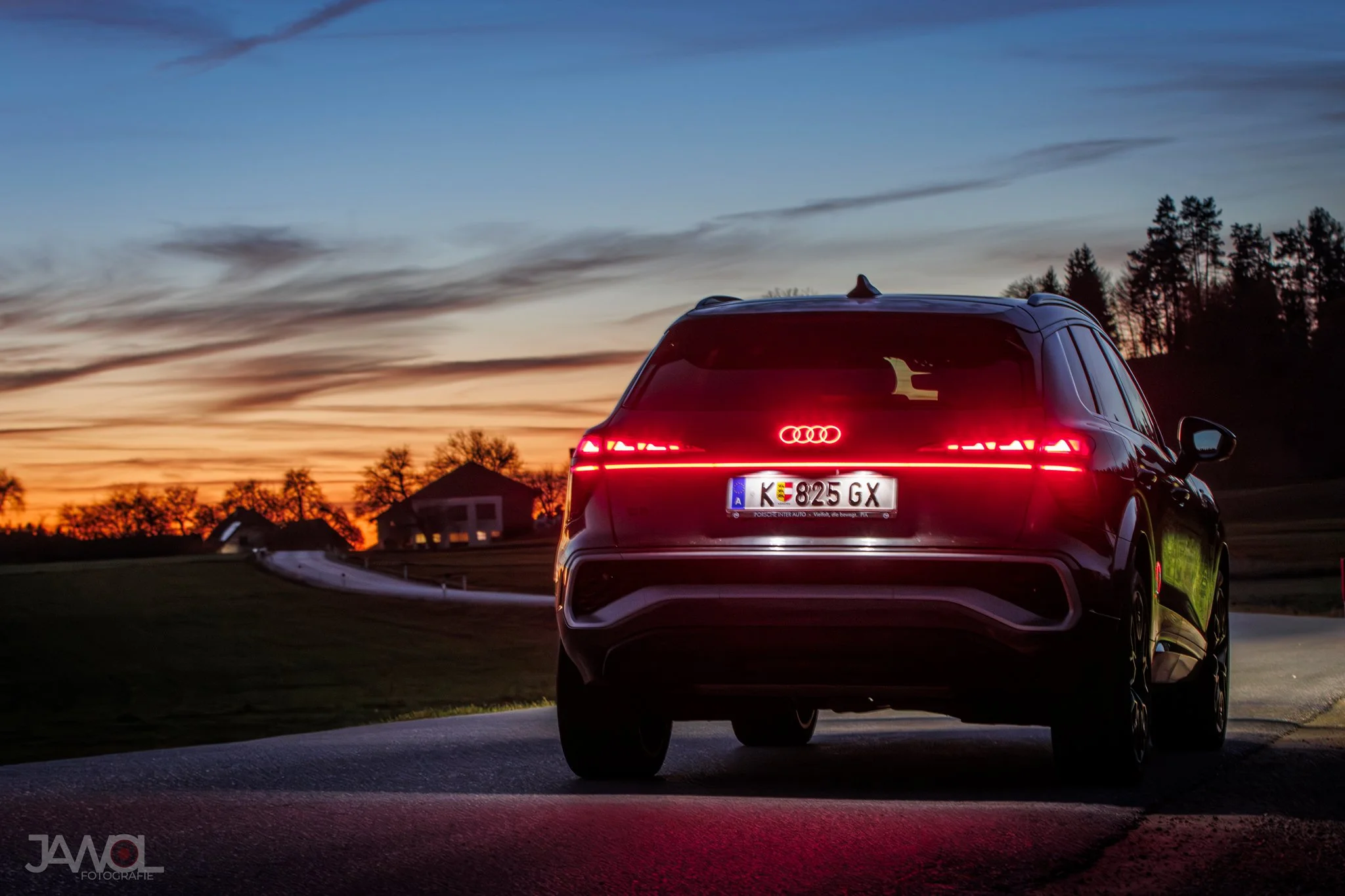 Ein schwarzer Audi SUV mit leuchtenden roten Rücklichtern auf einer Straße bei Sonnenuntergang, im Hintergrund Bäume und ein Haus.