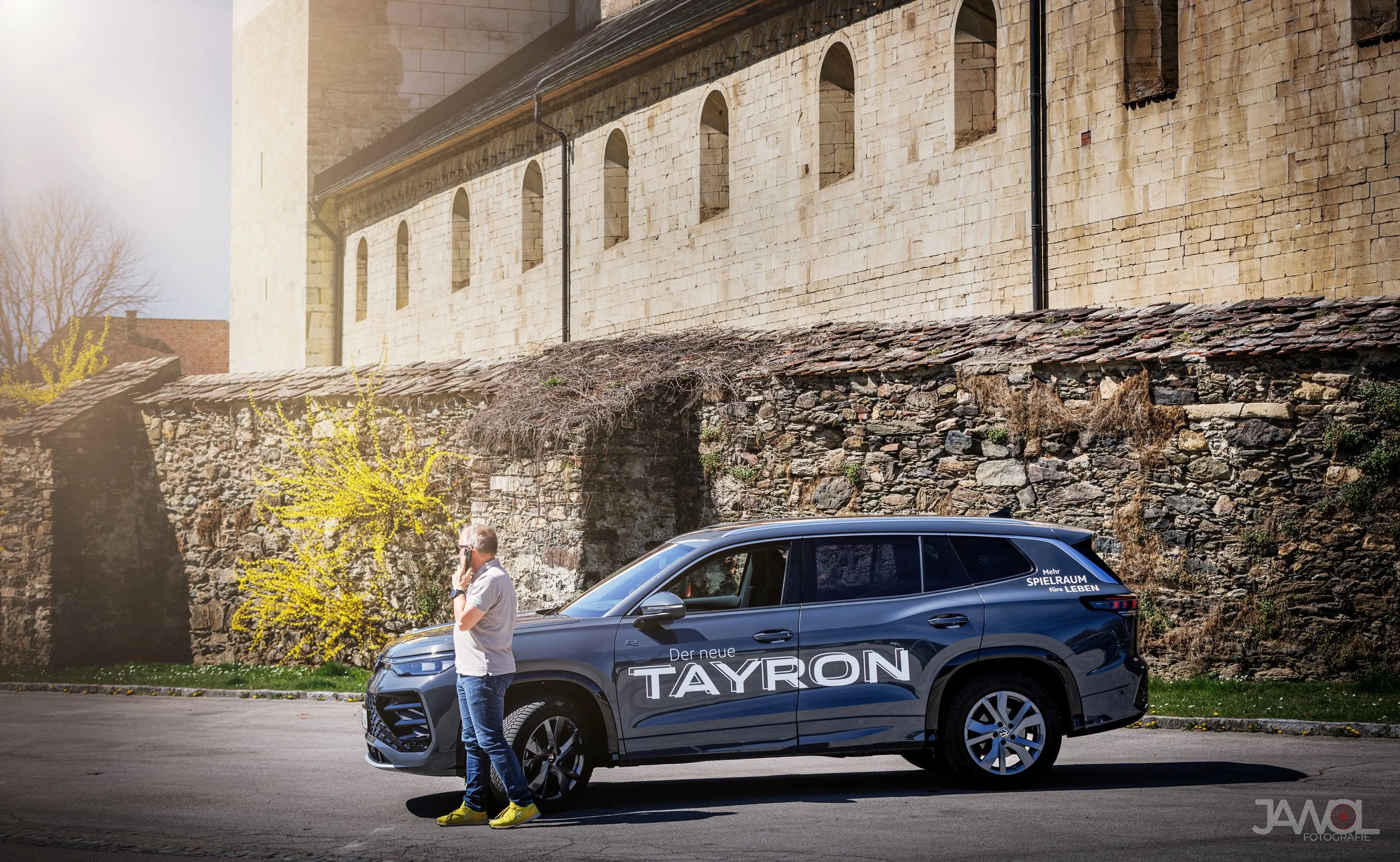 Ein Mann steht neben einem schwarzen SUV, das den Schriftzug "Der neue TAYRON2" trägt, während er telefoniert, im Hintergrund eine alte Steinmauer und ein historisches Gebäude.