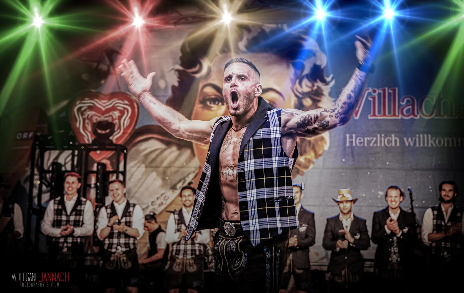 Mann mit Tattoo-Armen, lackiertem Oberkörper und offenem Kleidungsstück auf einer Bühne, mit offenen Armen, im Hintergrund eine Gruppe von Männern in Trachten und bunte Lichtstrahlen, hinter einem Wandbild einer Frau.