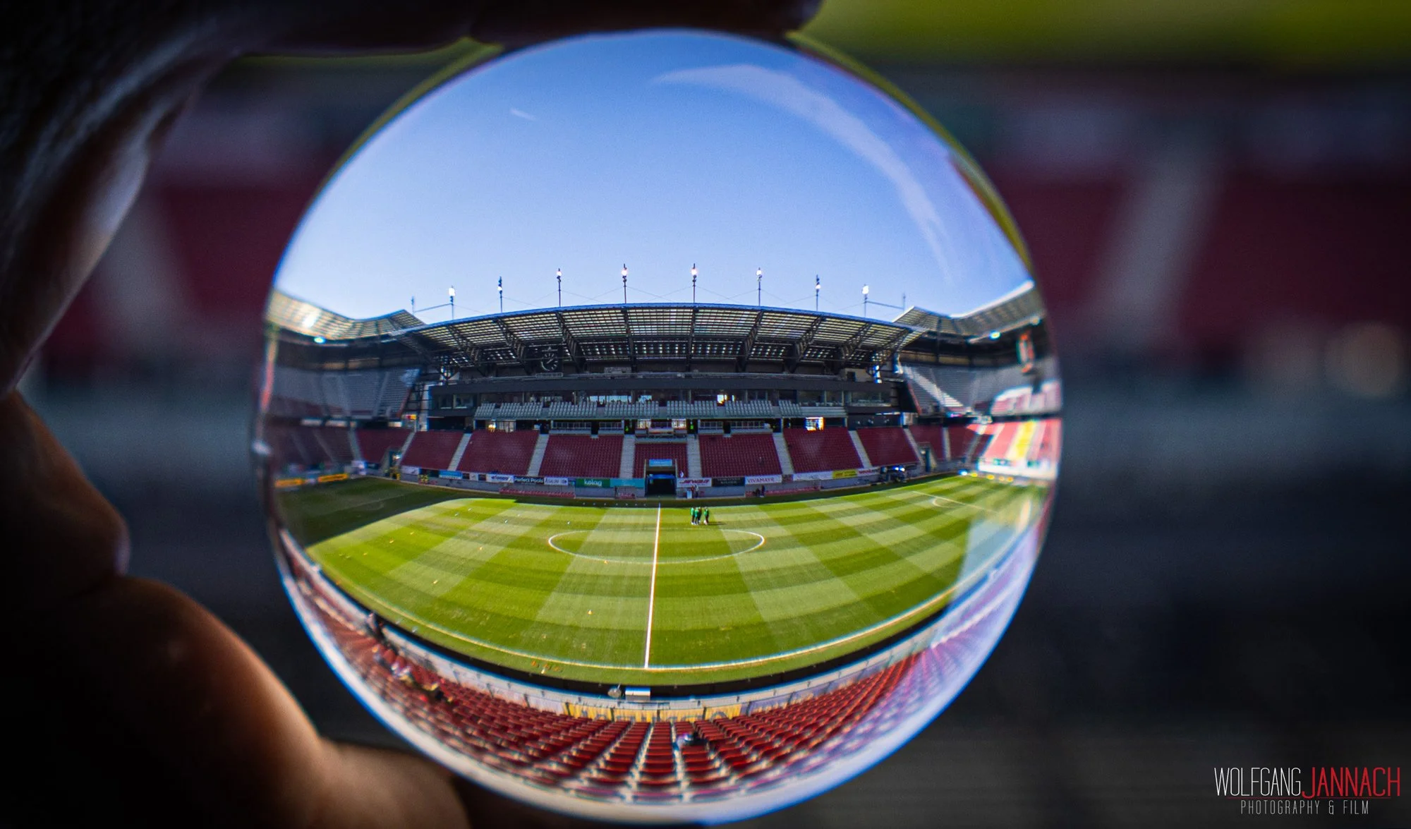 Leeres Fußballstadion mit grünem Rasen, Sicht durch eine Kristallkugel, blauer Himmel