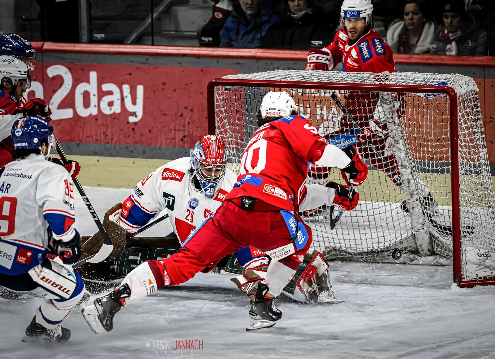 Eishockeyspieler in Aktion vor dem Tor, mit einem Torwart und einem Verteidiger, die versuchen, den Puck zu retten. Zuschauer im Hintergrund beobachten das Spiel.