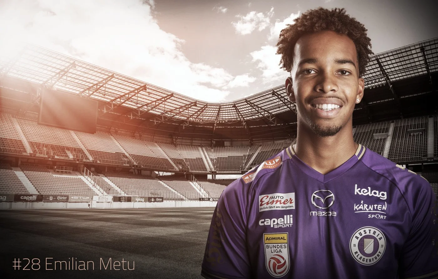 Porträt eines lächelnden Fußballspielers in lila Trikot im leeren Stadion, Text hit#28 Emilian Metu