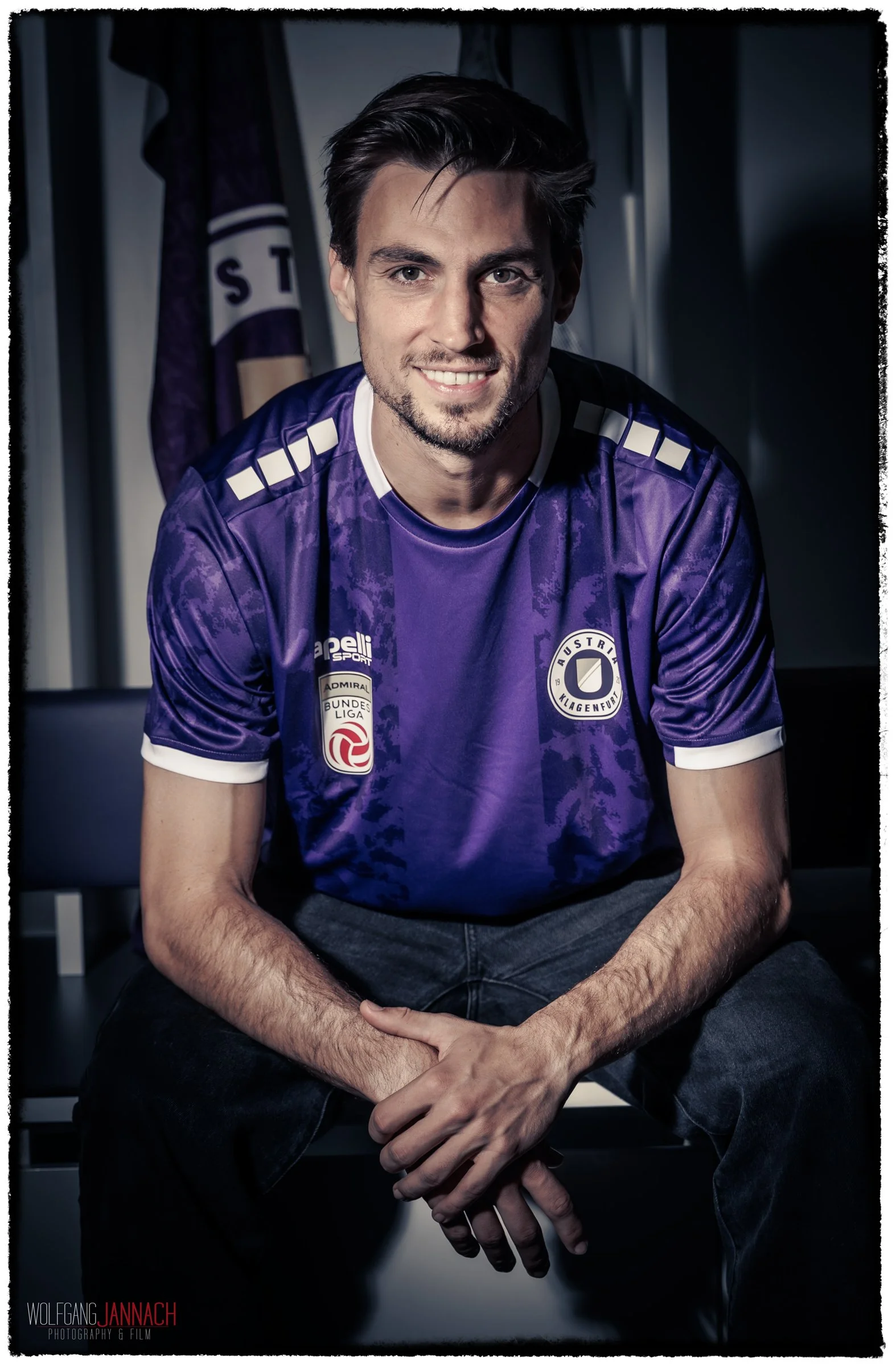 Mann in lila Trikot mit Logos, sitzend in einem Raum, lächelnd, dunkler Hintergrund.