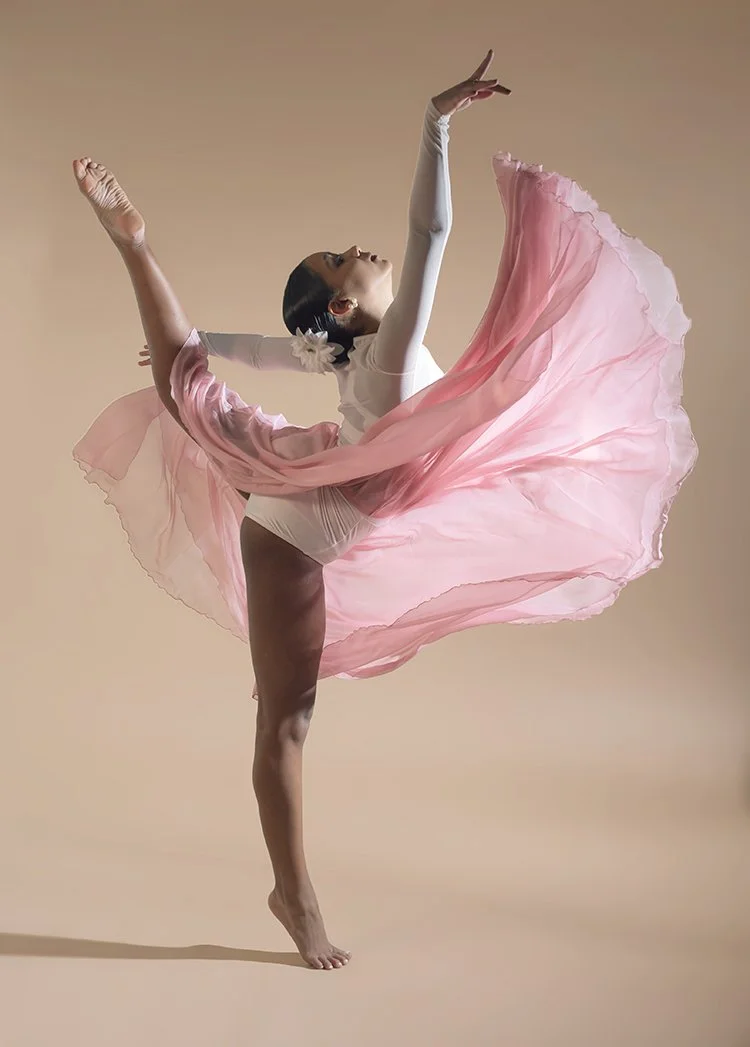 A ballet dancer in a pink flowing skirt and white top performing a high Arabesque pose against a plain beige background.