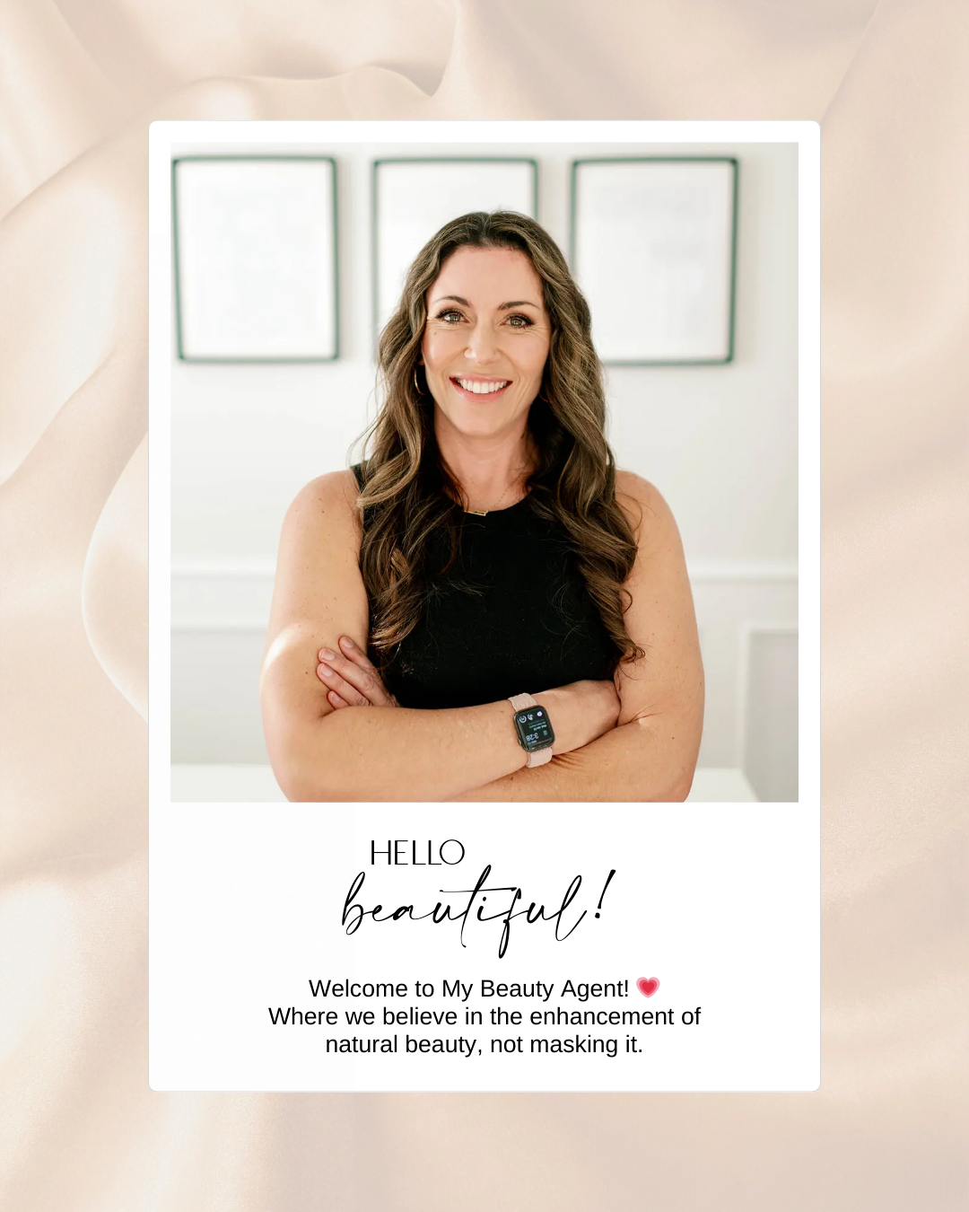 A woman with long, wavy brown hair smiling and standing with arms crossed, wearing a black sleeveless top and a smartwatch. Behind her are three framed pictures on a white wall.