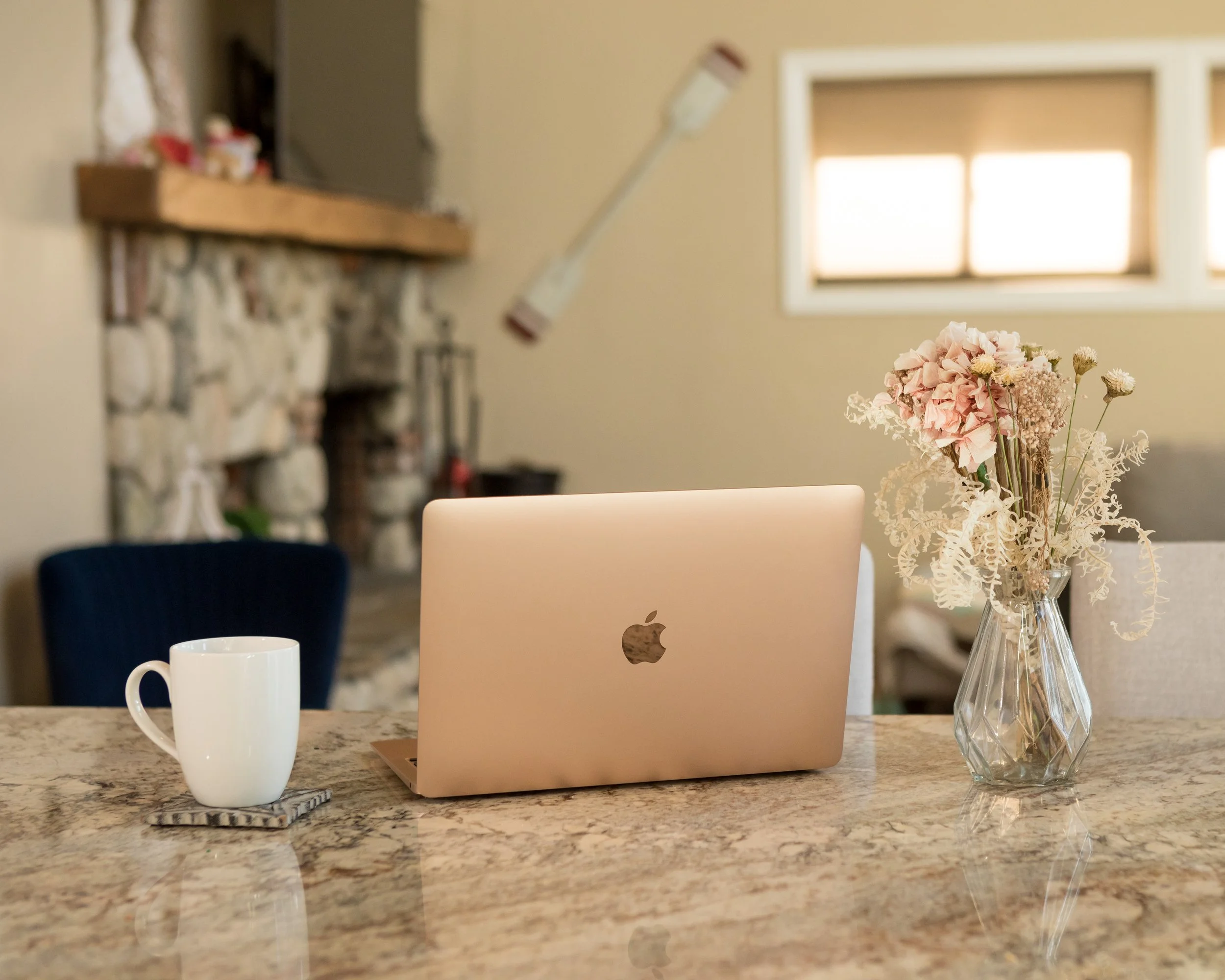 A kitchen countertop with a white coffee mug, a closed laptop, and a clear glass vase with pink and white flowers. In the background, there is a stone fireplace, a window with sunlight coming through, and a bouquet of dried flowers.