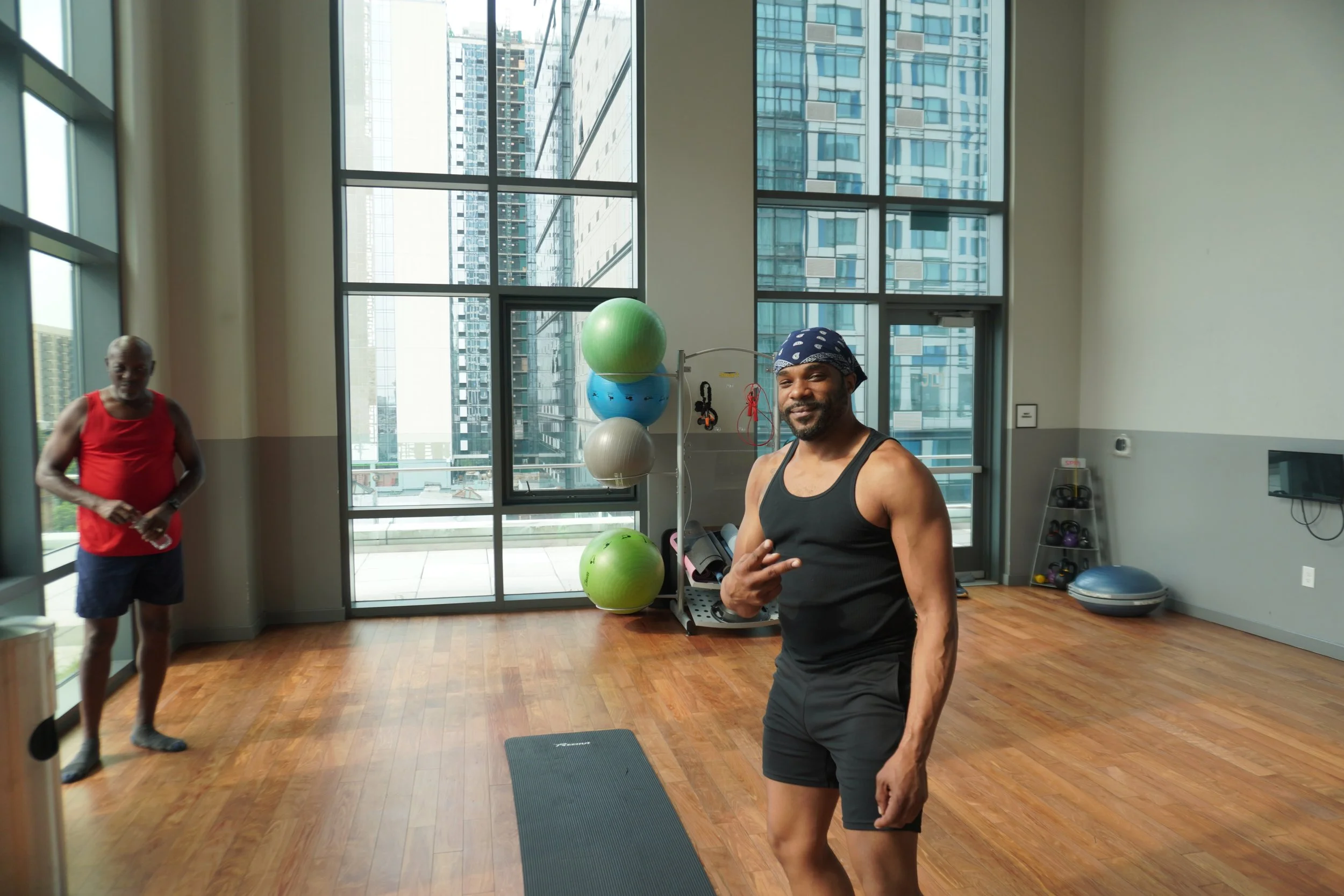 Two men in workout clothes in a gym with large windows and city view. One man is smiling and posing, the other is standing in the background.