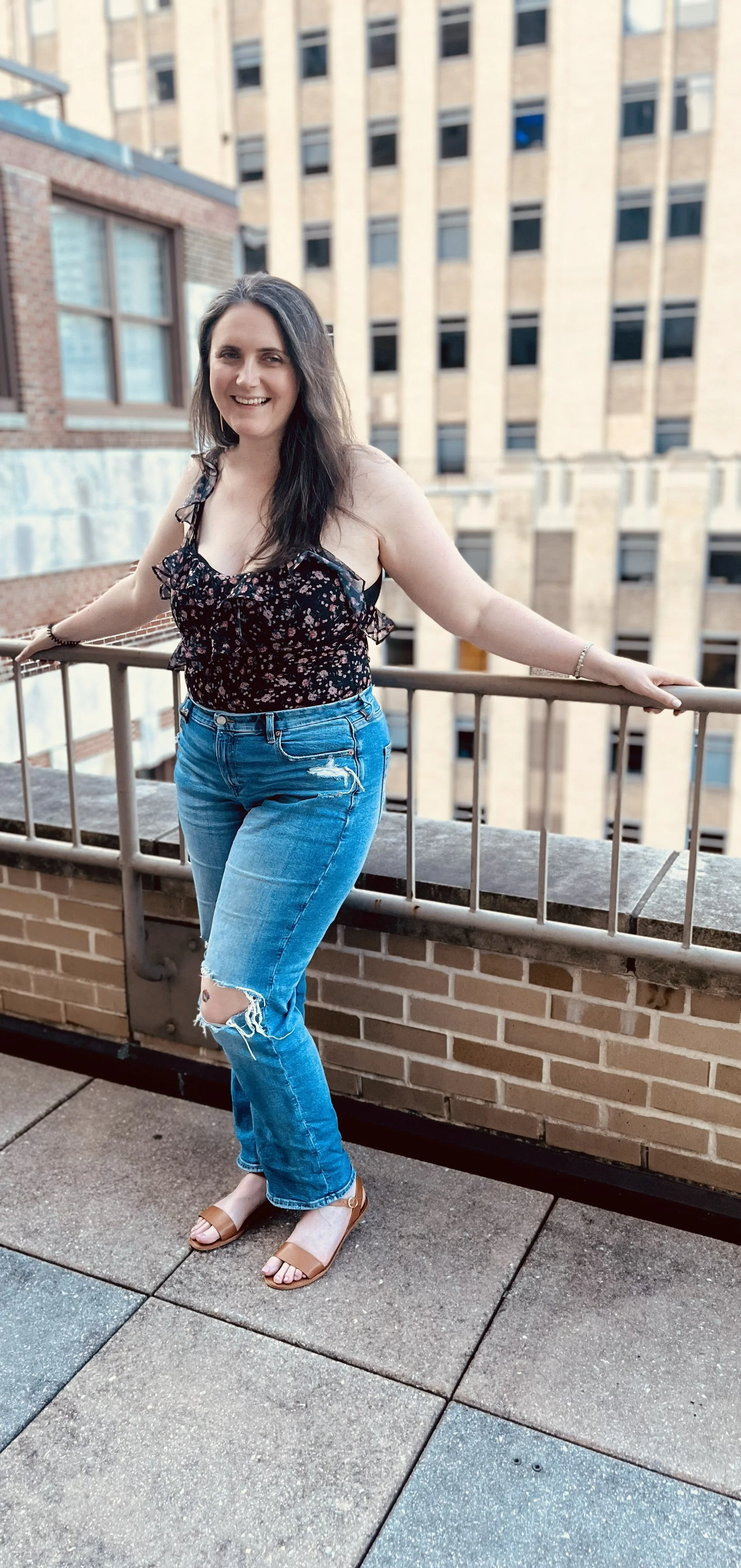 A woman with long dark hair smiling and standing on a rooftop balcony with a cityscape of tall buildings behind her. She is wearing a floral sleeveless top, ripped blue jeans, and brown open-toe sandals.