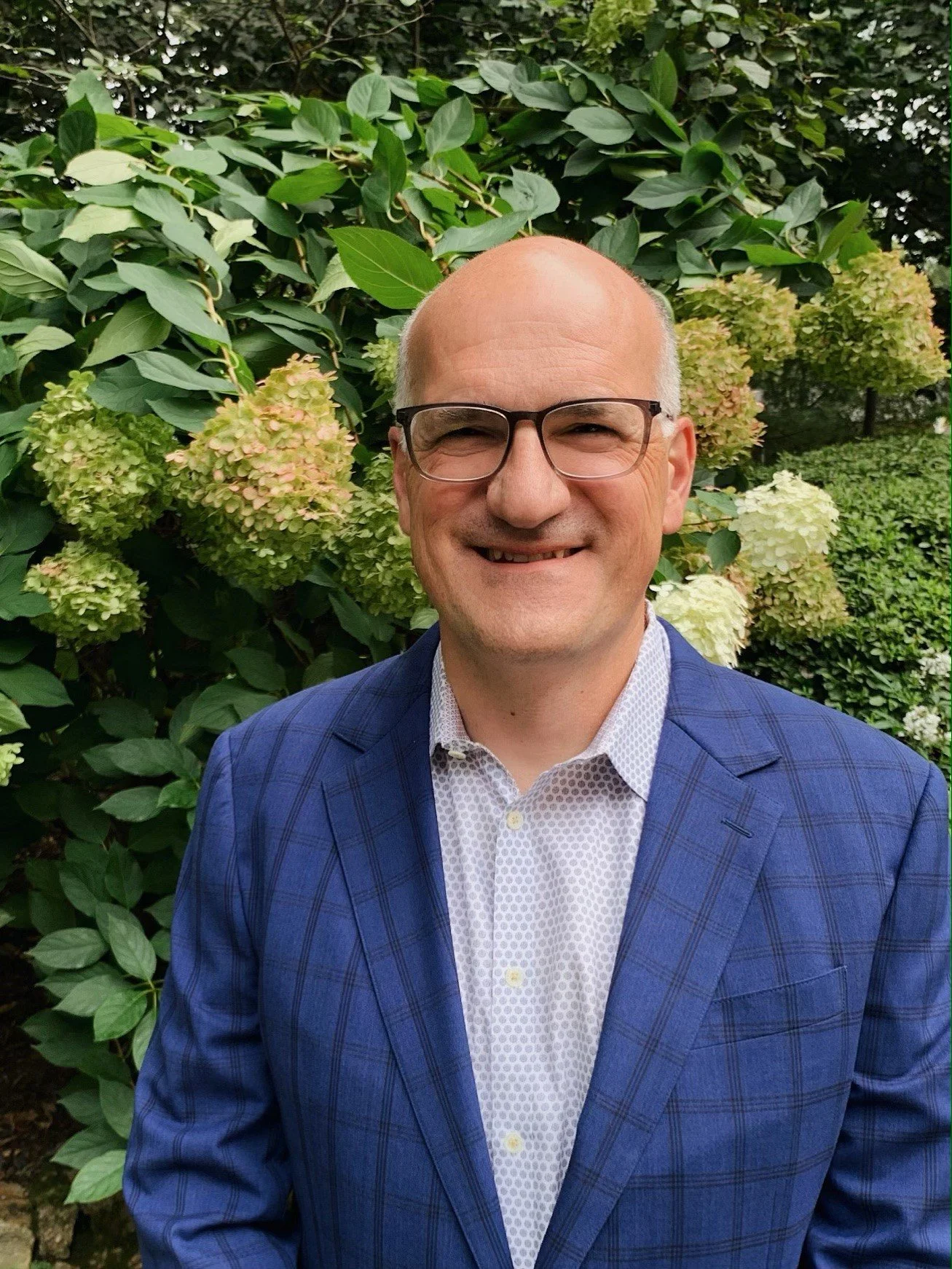 A smiling middle-aged man with glasses, wearing a blue plaid blazer and a white patterned shirt, standing in front of a bush with large, light-colored hydrangea flowers.