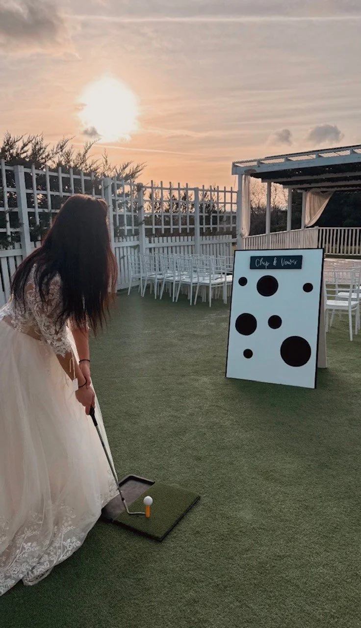 A woman in a wedding dress playing mini-golf on a grassy outdoor course during sunset, with a white fence and a black and white cornhole board in the background.