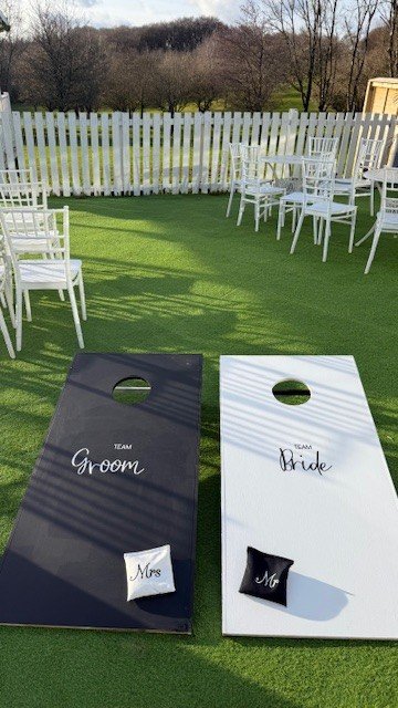 Cornhole boards labeled for the groom and bride with small pillows marked 'Mrs.' and 'Mr.' in front of white chairs on a lawn.