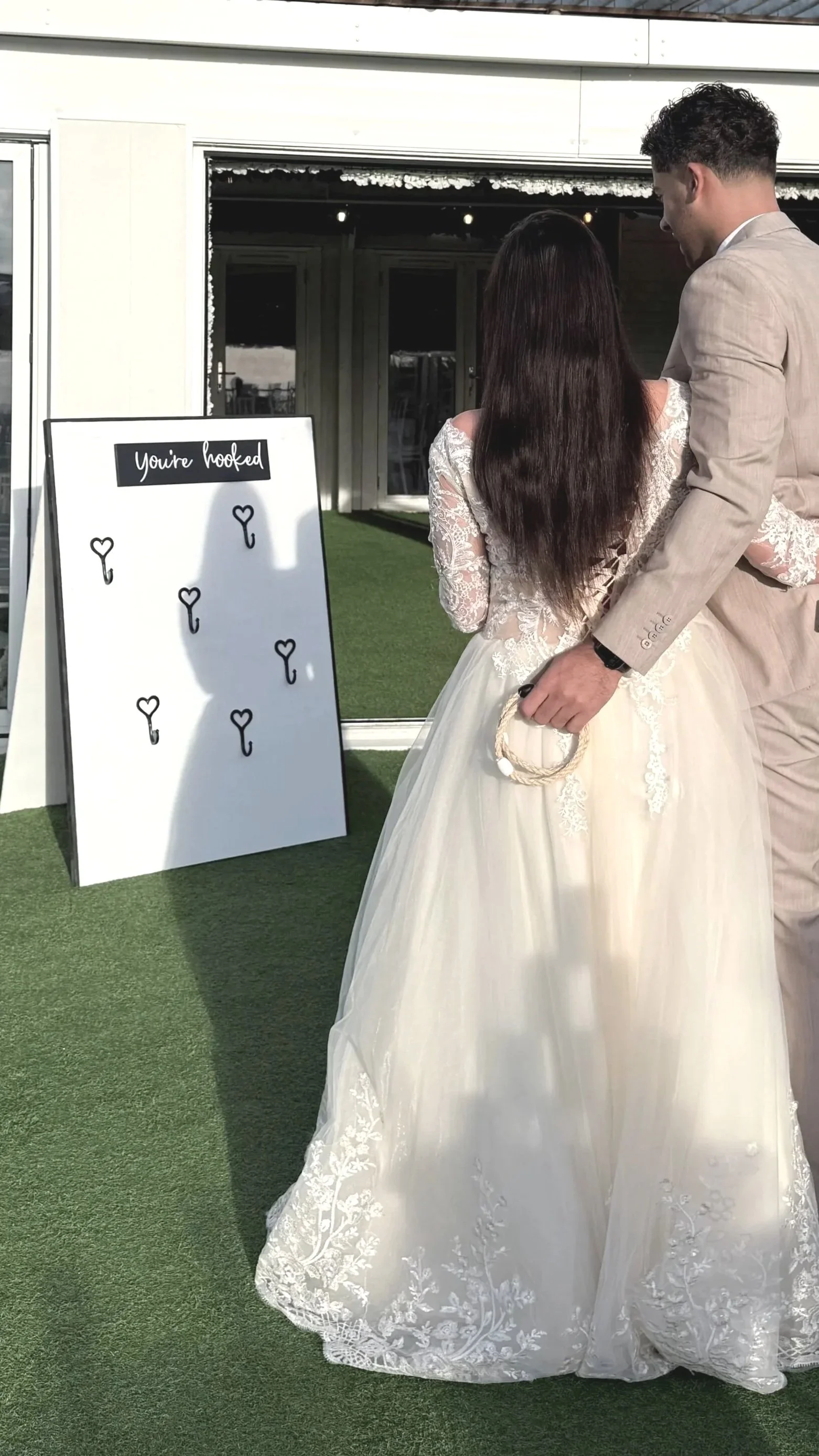 A bride and groom standing outside in front of a white board with the words "You're hooked" written at the top, with fishing hook illustrations. The bride is wearing a white lace wedding dress, and the groom is in a beige suit.
