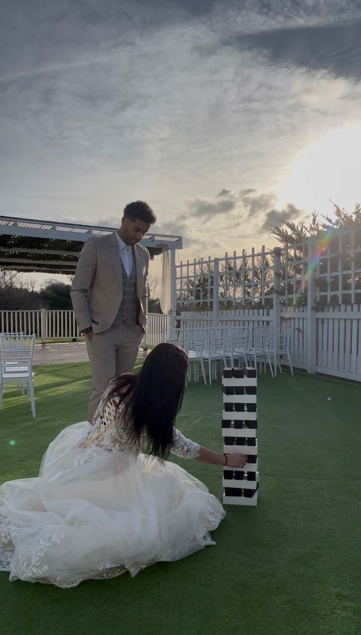 A man in a beige suit and a woman in a white wedding dress are playing Jenga outdoors on the grass during sunset. The woman is kneeling and removing a block from the Jenga tower, while the man stands watching.