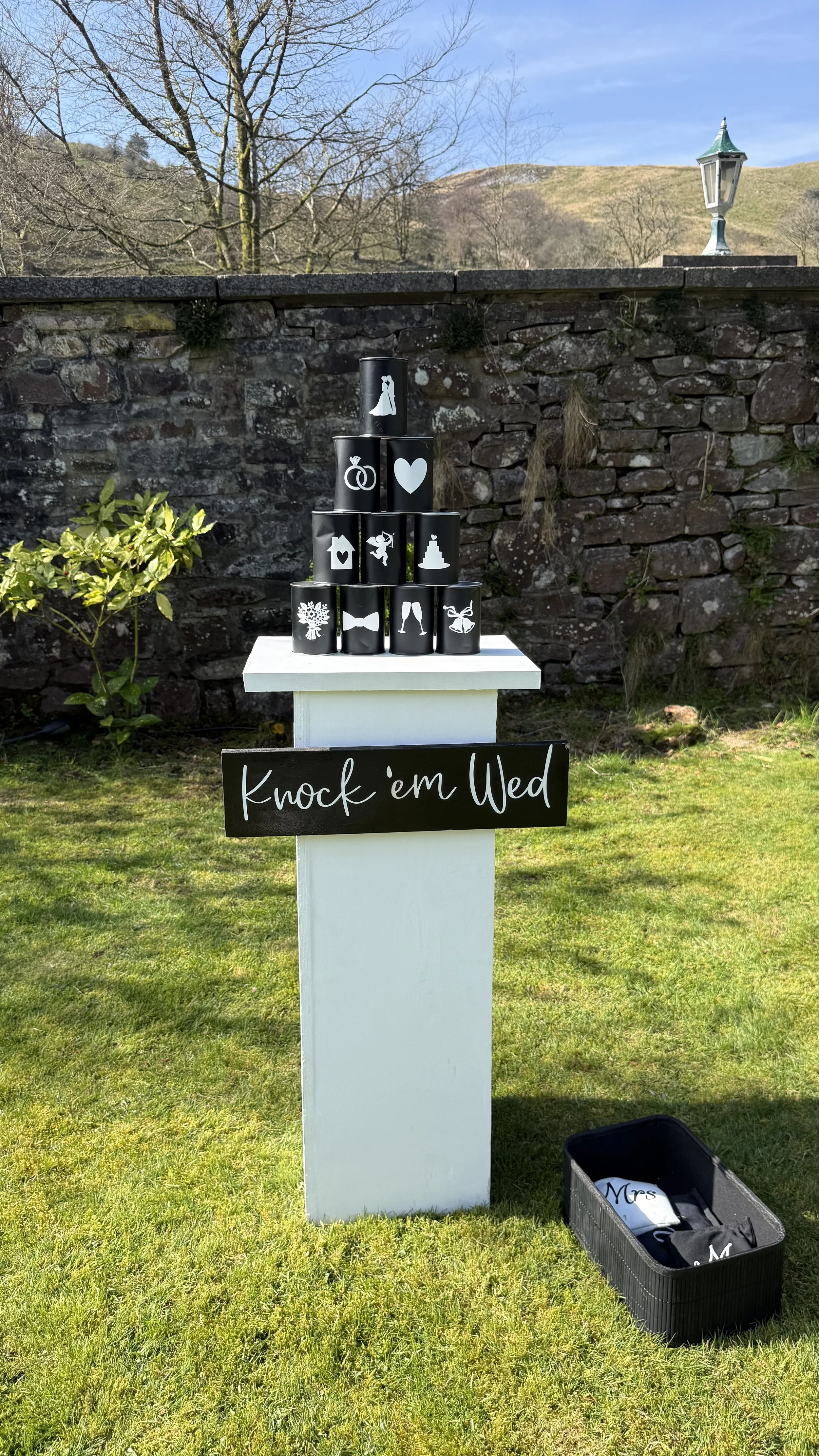 Outdoor wedding decoration featuring a small display with black cans decorated with wedding-themed icons, stacked in a pyramid on a white stand. A black sign with white handwritten text reads 'Knock 'em Wed'. A basket containing personalized items is