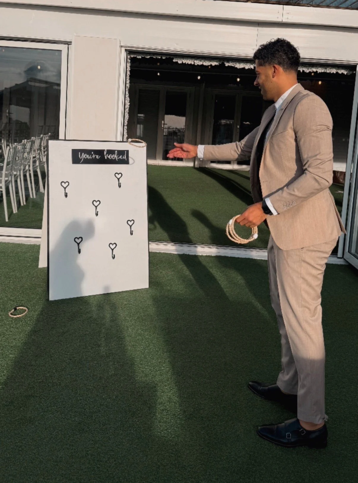 A man in a beige suit is playing a game of cornhole outdoors at sunset, aiming to toss rings onto a board with the words 'You're hooked' and black heart sketches.