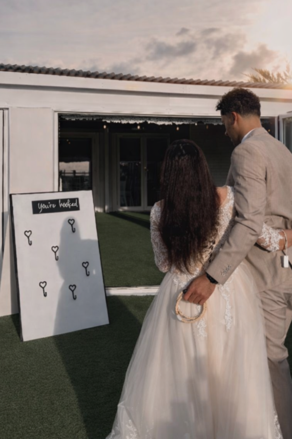 A bride and groom stand together on a green lawn, looking at a large white board with black markers and the words 'you're booked' written on it, during a wedding event at sunset.