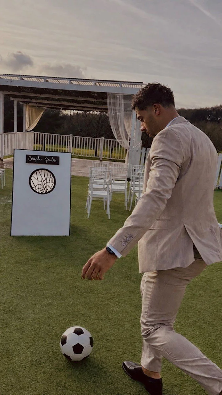 A man in a beige suit playing with a soccer ball outdoors near a wedding setup with white chairs and drapes.