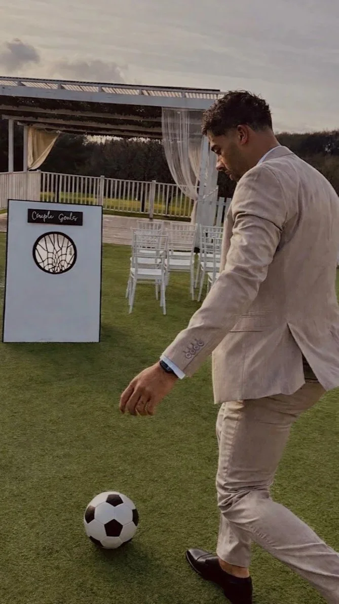 Man in a beige suit playing soccer on a grassy outdoor area, with a cornhole game and white chairs in the background.