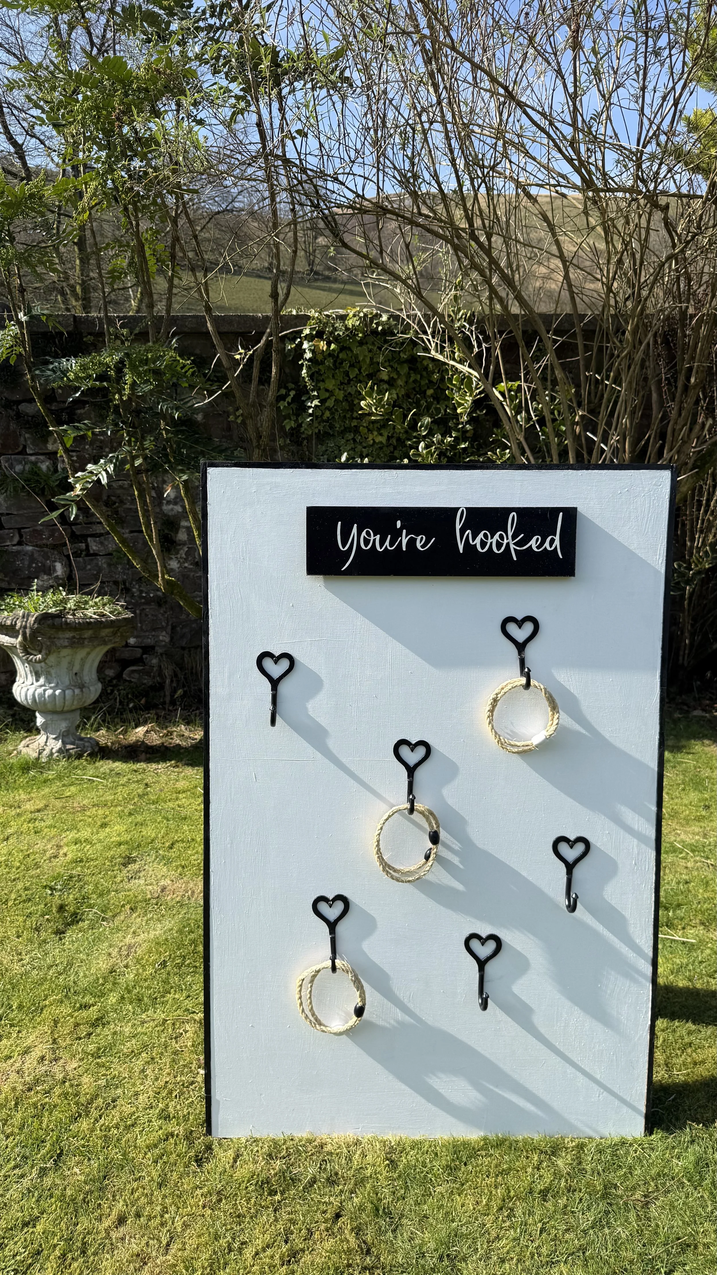 A game board with the phrase 'You're hooked' at the top and six hooks attached to a white board with black hearts as handles; some hooks hold decorative rings, while others are empty, set outdoors on a grassy lawn.