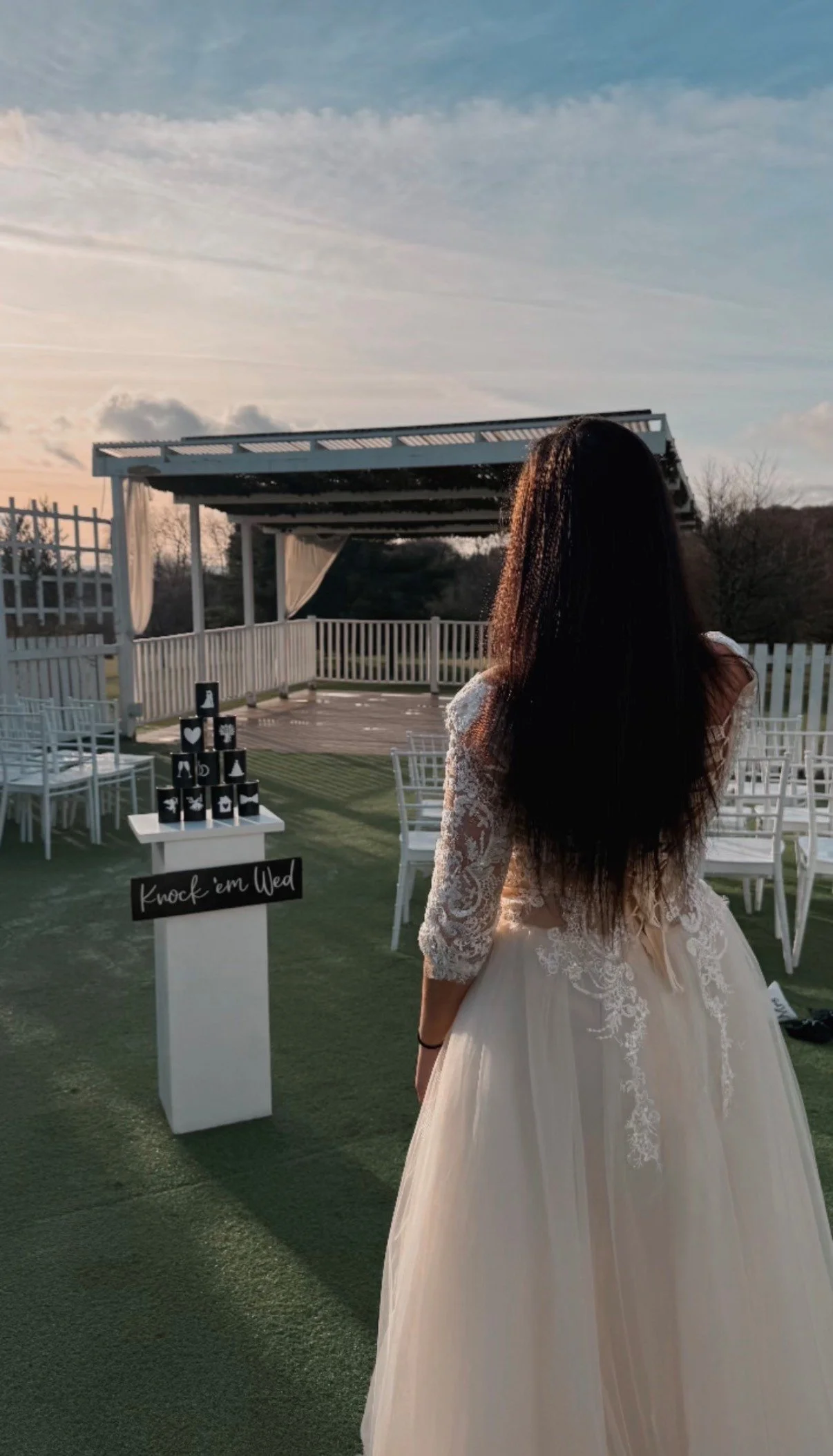 A woman in a wedding dress standing outdoors at a wedding venue with a small stage, white chairs, and a sign that says 'Knock 'em Dead' during sunset.