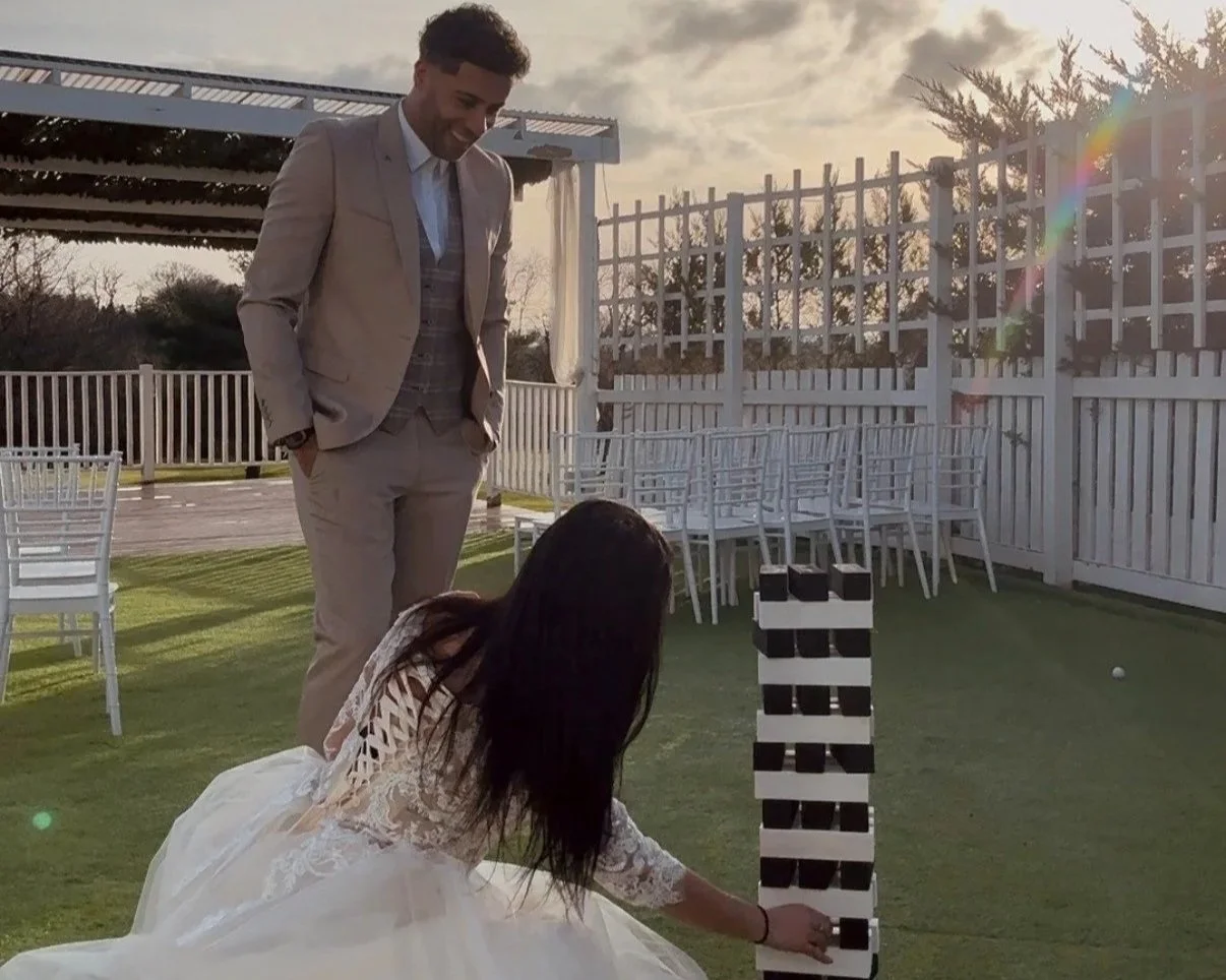 A man in a beige suit and a woman in a white lace dress playing Jenga outdoors on a grassy area with white chairs and fence, during sunset.