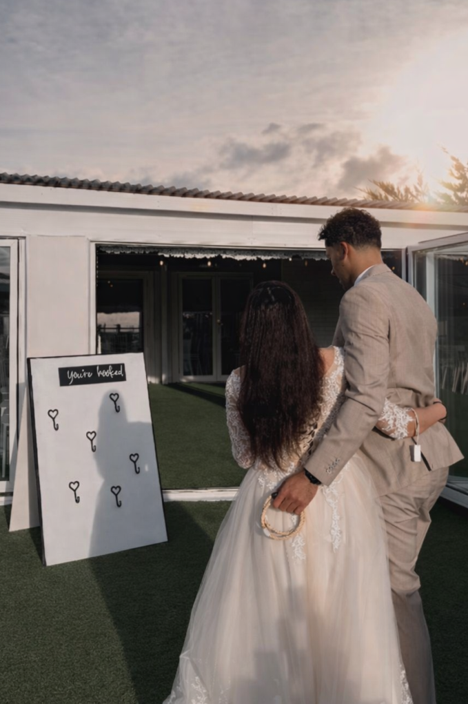 A couple dressed in wedding attire, the woman in a white lace wedding gown and the man in a beige suit, embrace outdoors near a house with an open sliding door. A sign with the words "you're hooked" and small key illustrations is visible nearby, with a sunset sky in the background.