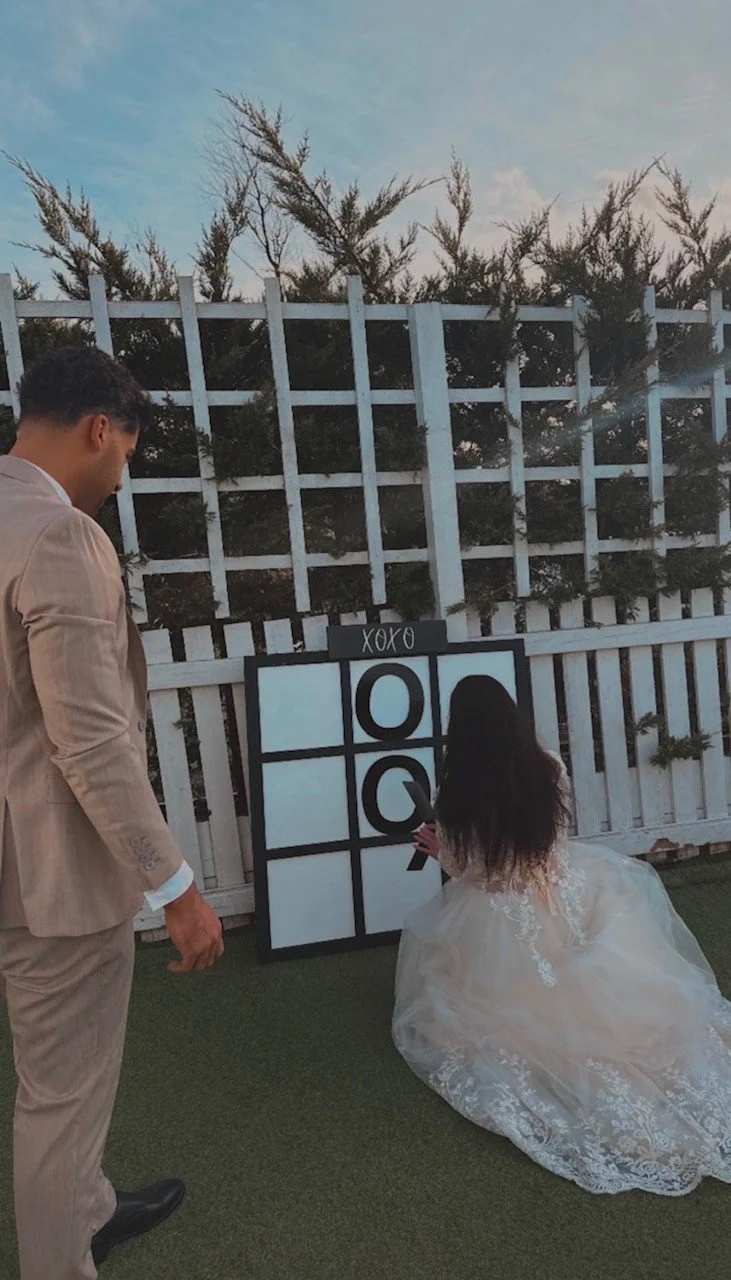 A woman in a wedding dress is playing tic-tac-toe on a large board outdoors, while a man in a beige suit watches.