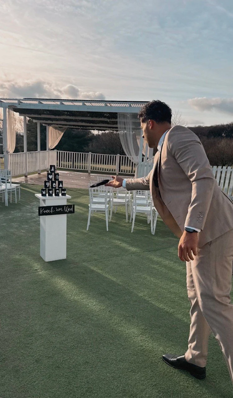 A man in a beige suit is playing a game of knock 'em down with stacked black cans on a table outside on a decorated patio, with a background of a white fence, white chairs, and a pergola under a partly cloudy sky.