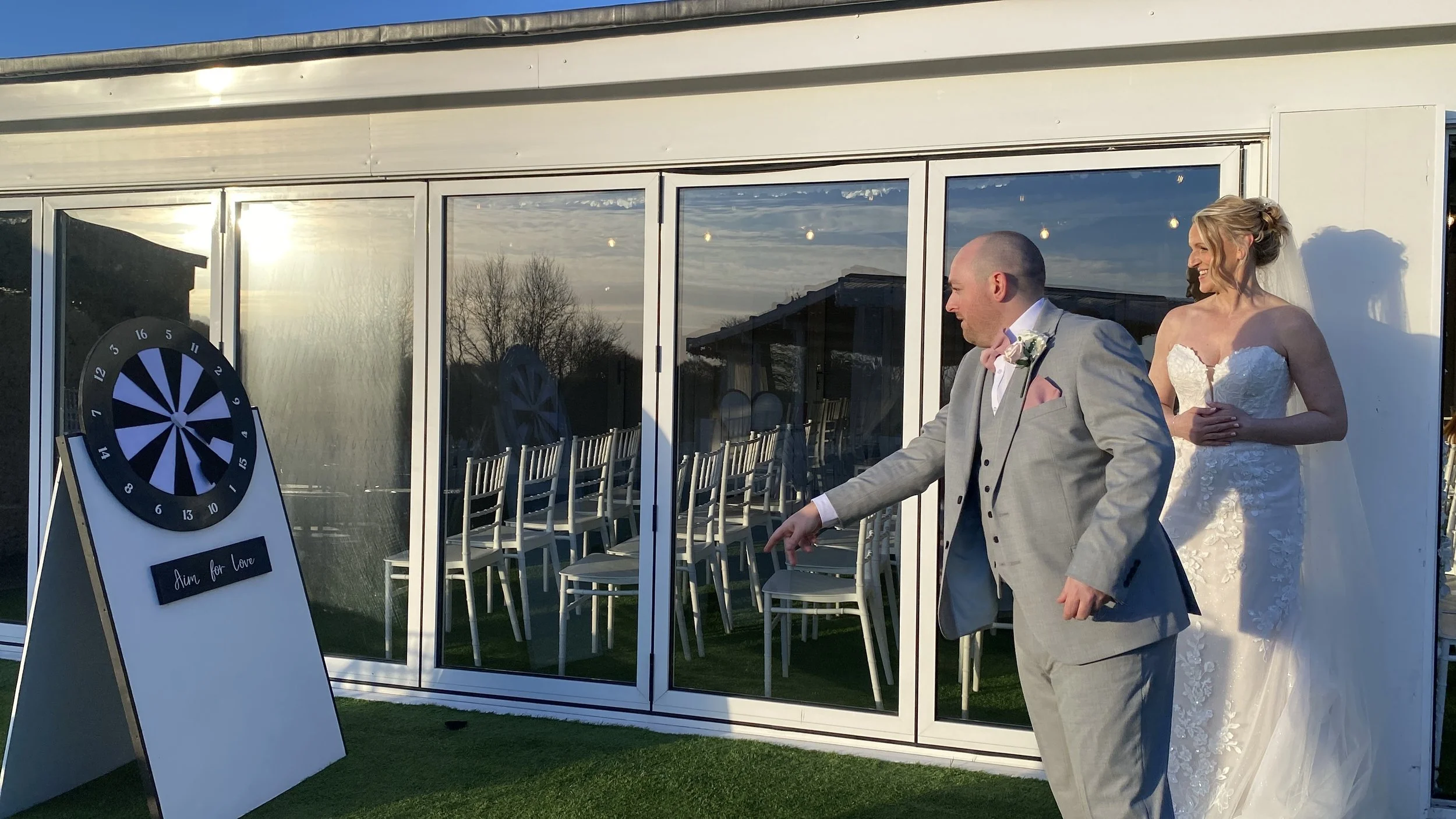 A wedding scene with a bride and groom playing darts outside. The bride in a white lace wedding dress stands behind the groom, who is in a light gray suit. The scene is lit by the sunset, and inside the glass-walled building, empty chairs are visible