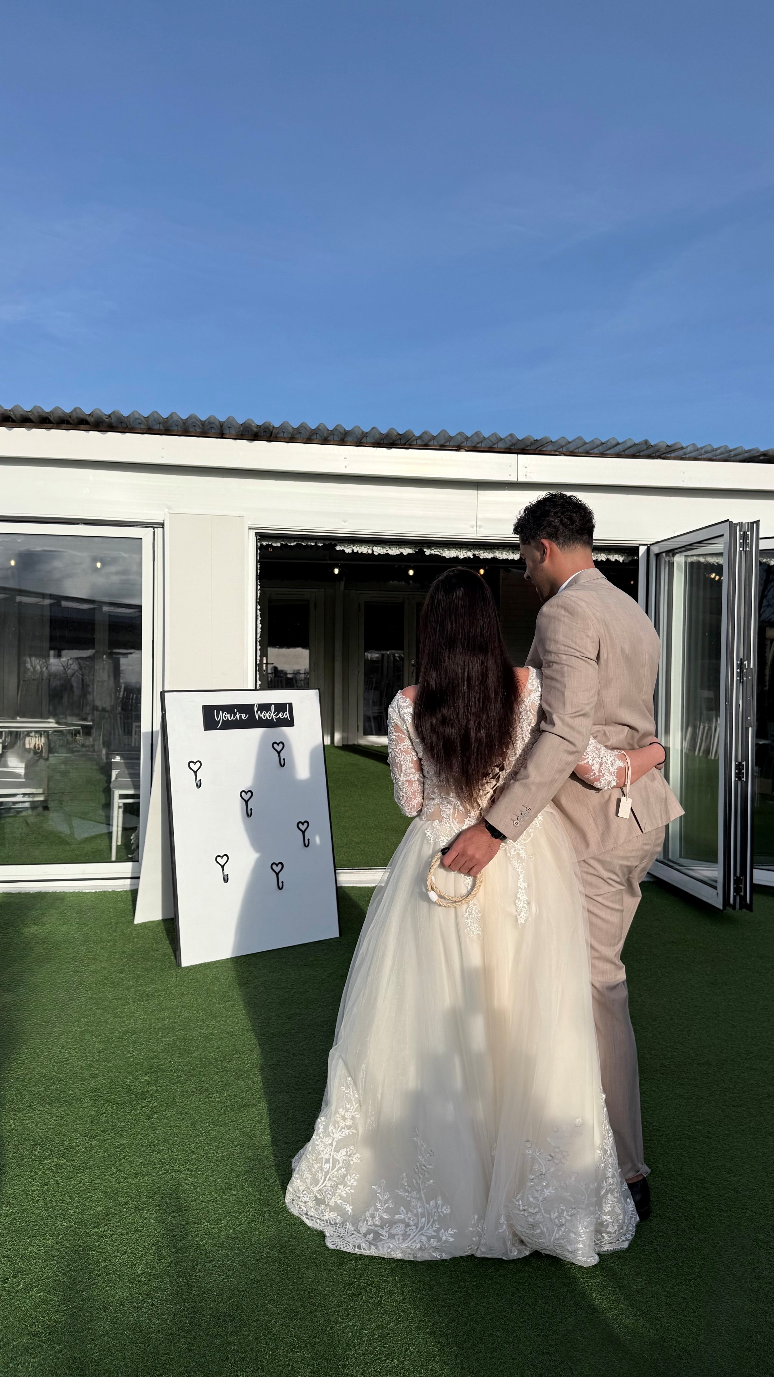 A bride and groom, dressed in wedding attire, hugging each other outdoors near a white building with glass doors. A whiteboard sign with black text that reads 'You're hooked' stands nearby, decorated with small black key illustrations.