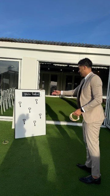 A man in a suit about to toss a ring onto a hook on a white board at an outdoor event during daytime.