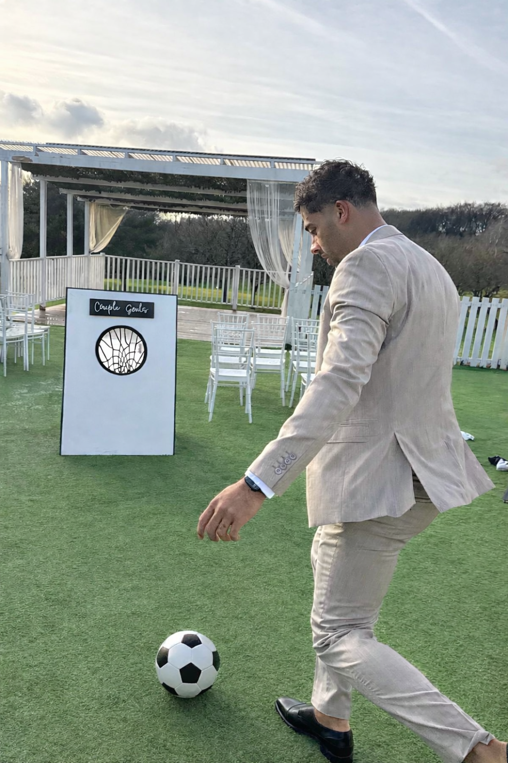 A man in a beige suit playing soccer on an outdoor artificial grass field at a wedding or event venue, with chairs and a white decorated canopy in the background.