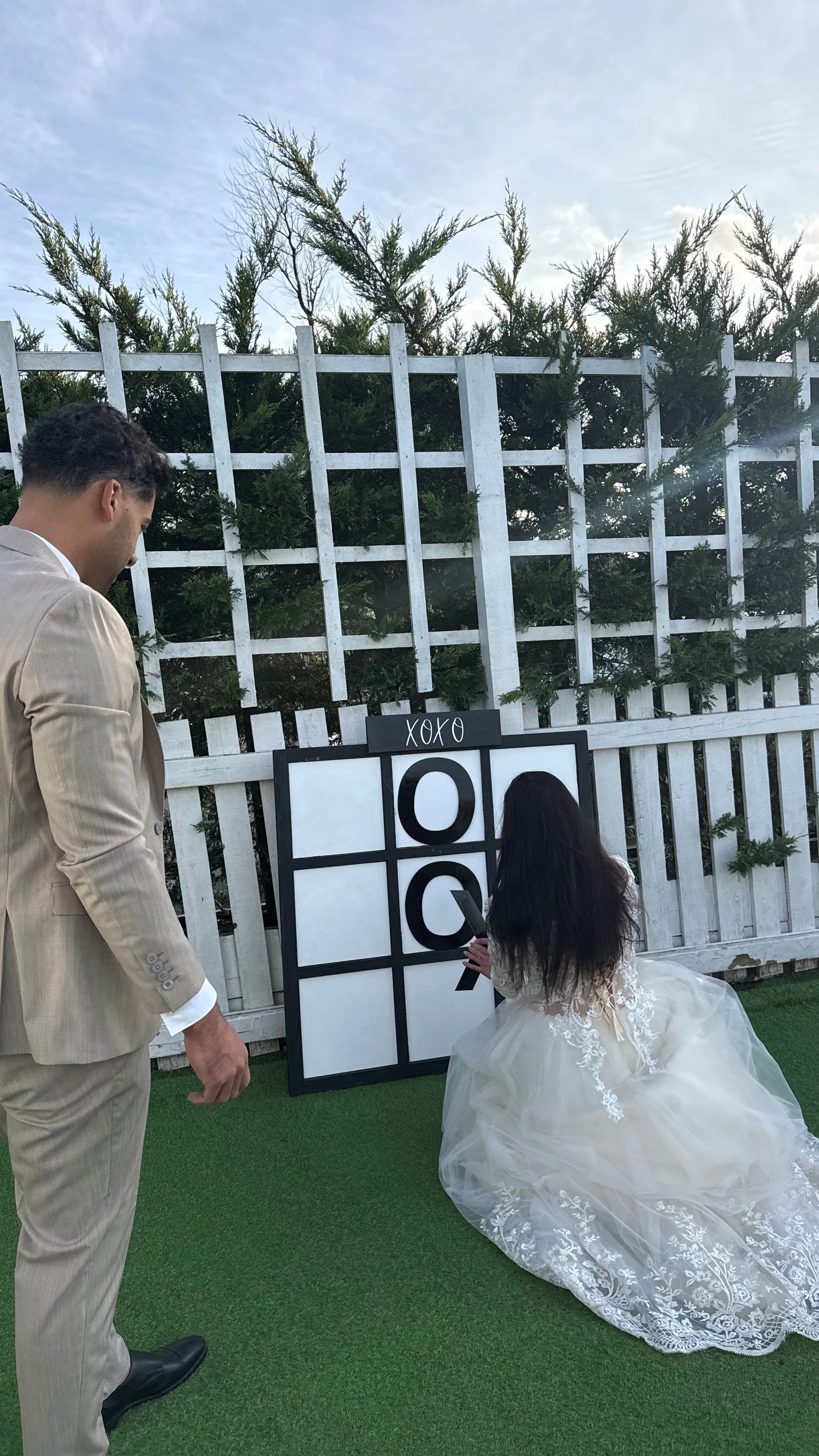 A man in a beige suit and a woman in a white wedding dress playing tic-tac-toe outdoors on a large black-and-white board, with a white fence and greenery in the background.