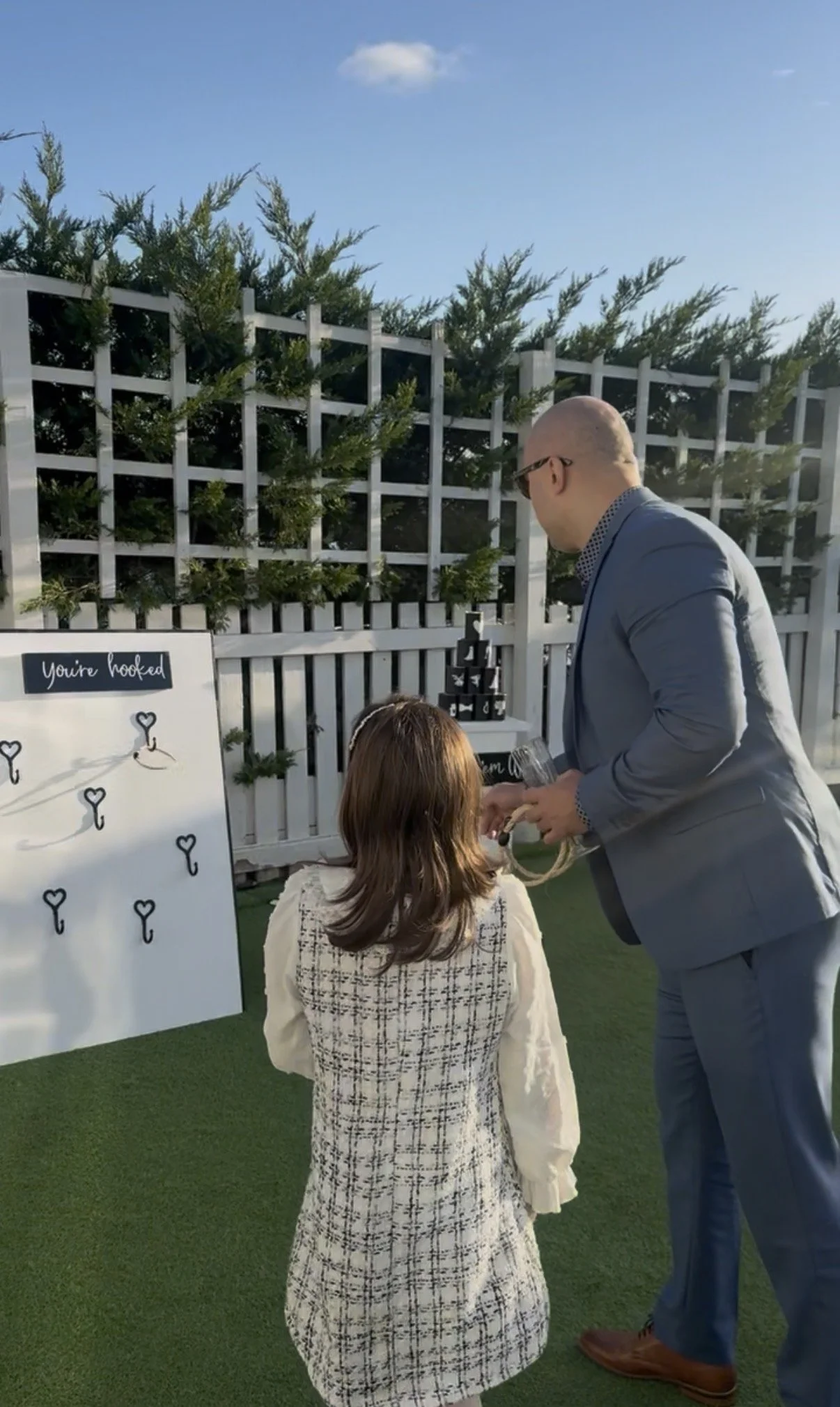 A man in a blue suit and a young girl are at an outdoor event, with a white wooden lattice fence decorated with greenery behind them. The man appears to be handing something to the girl near a game or activity involving small keys on a whiteboard lab
