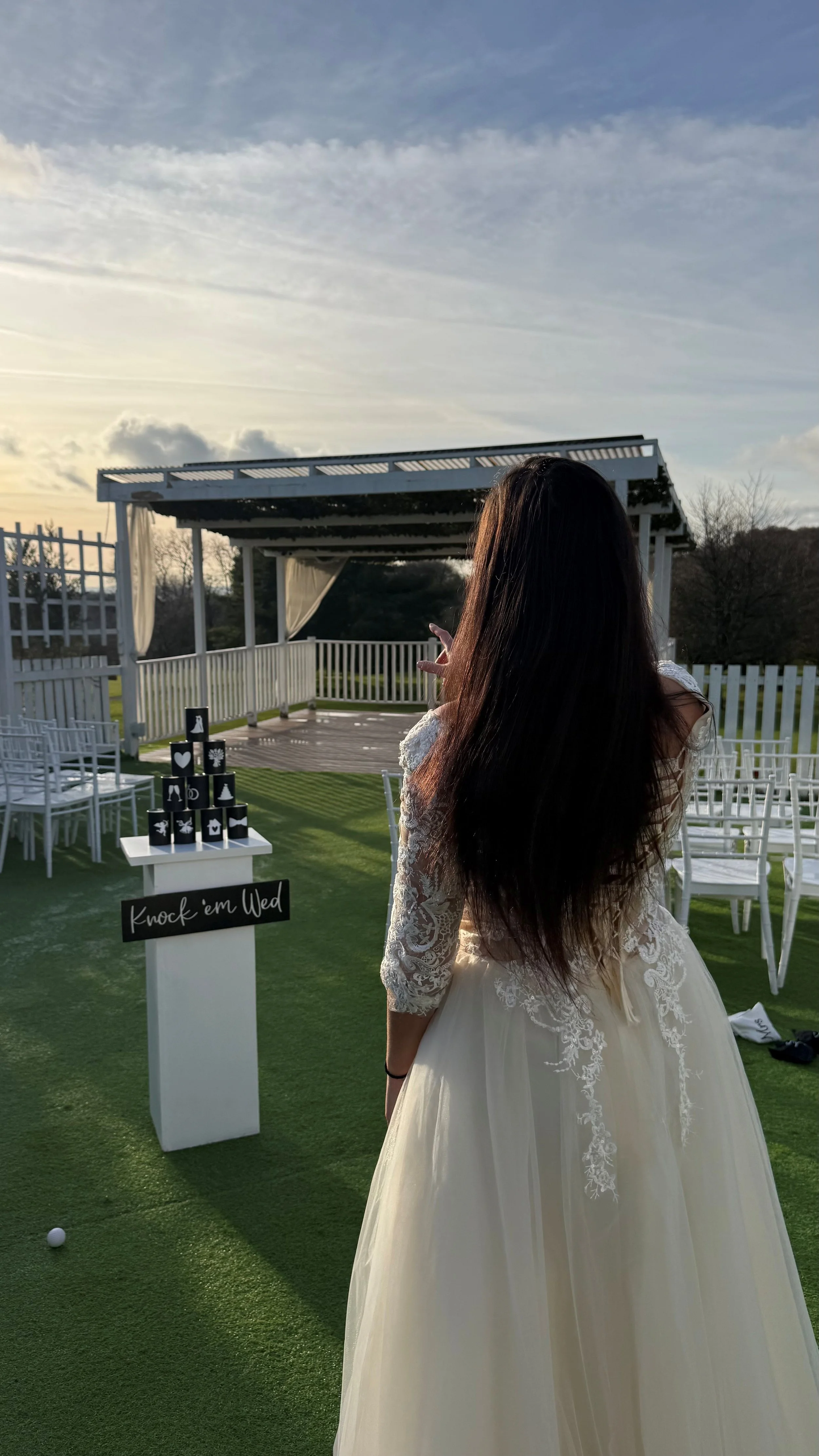 A woman in a white lace wedding dress stands outdoors at sunset, pointing towards a stack of black cups with white designs on a table labeled 'Knock 'em Wed'. There are white chairs, a small stage, and a white fence in the background, with a grassy a
