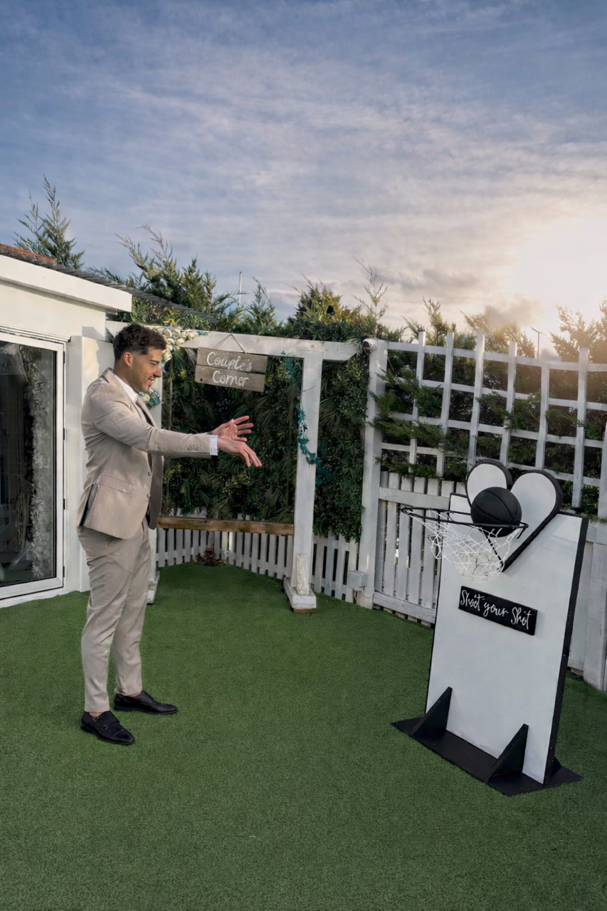 Young man in a beige suit playing basketball in a backyard with a small basketball hoop and backboard that reads 'Shoot your Shot,' with a sunny sky and greenery in the background.