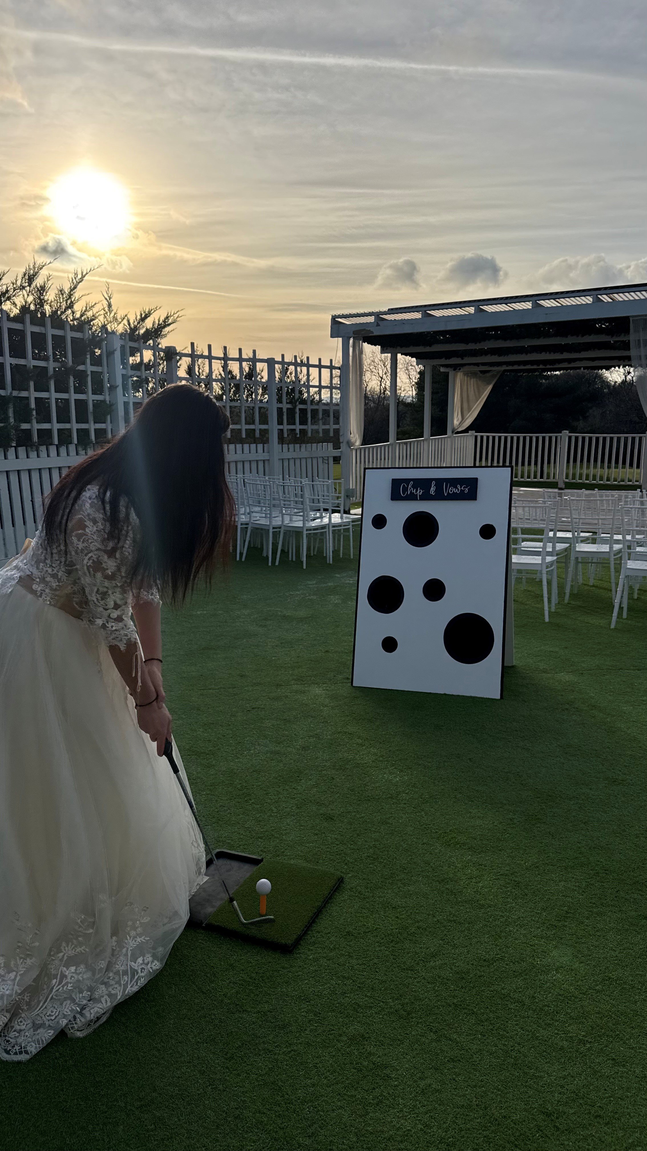 Woman in a wedding dress preparing to hit a golf ball on a mini golf course during sunset, with a large mockup of a game of chess in the background.