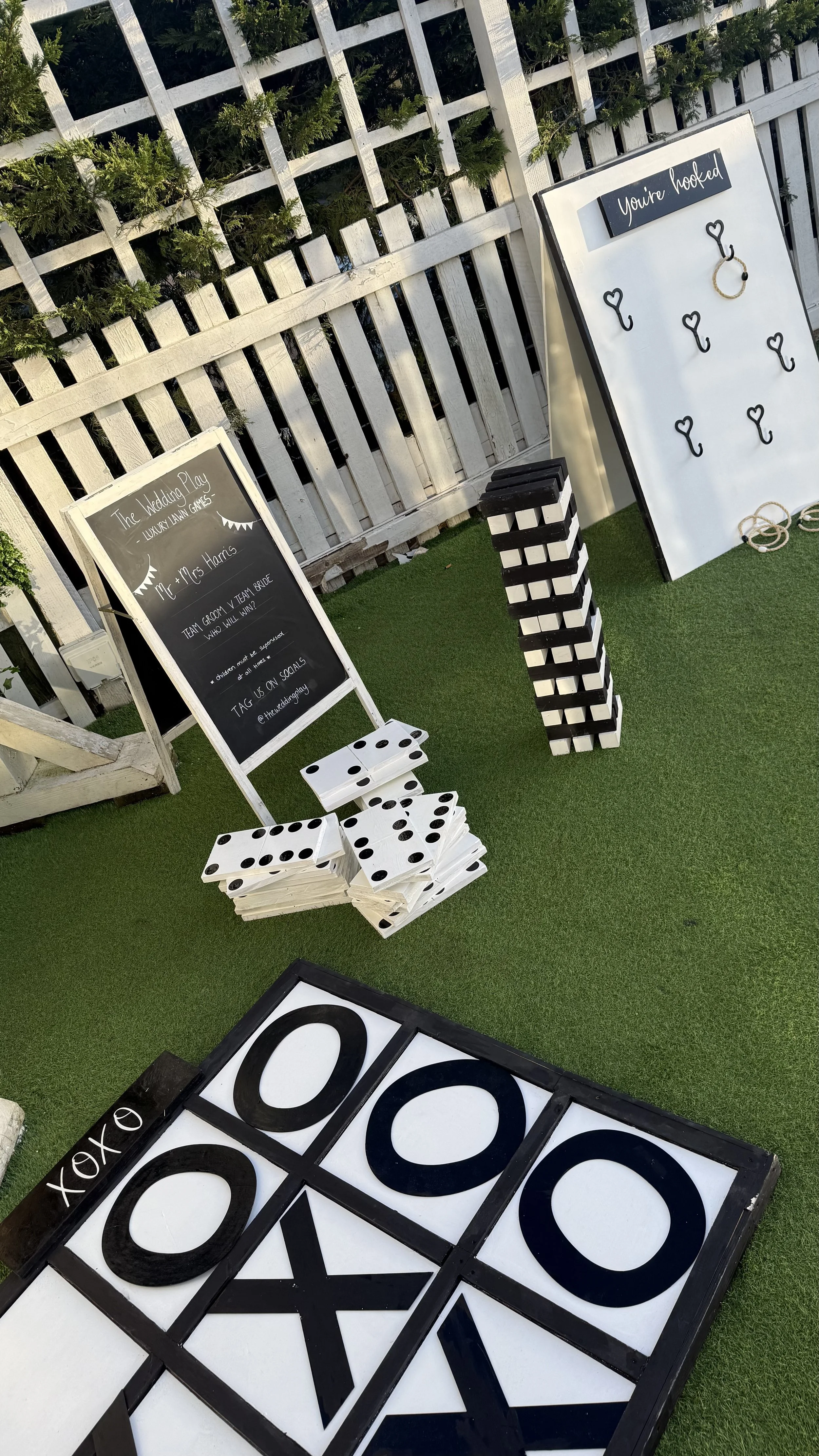 Setup for a wedding-themed outdoor game area featuring large domino and tic-tac-toe boards, with signs indicating it is part of wedding play activities, and decor including a white fence with greenery.