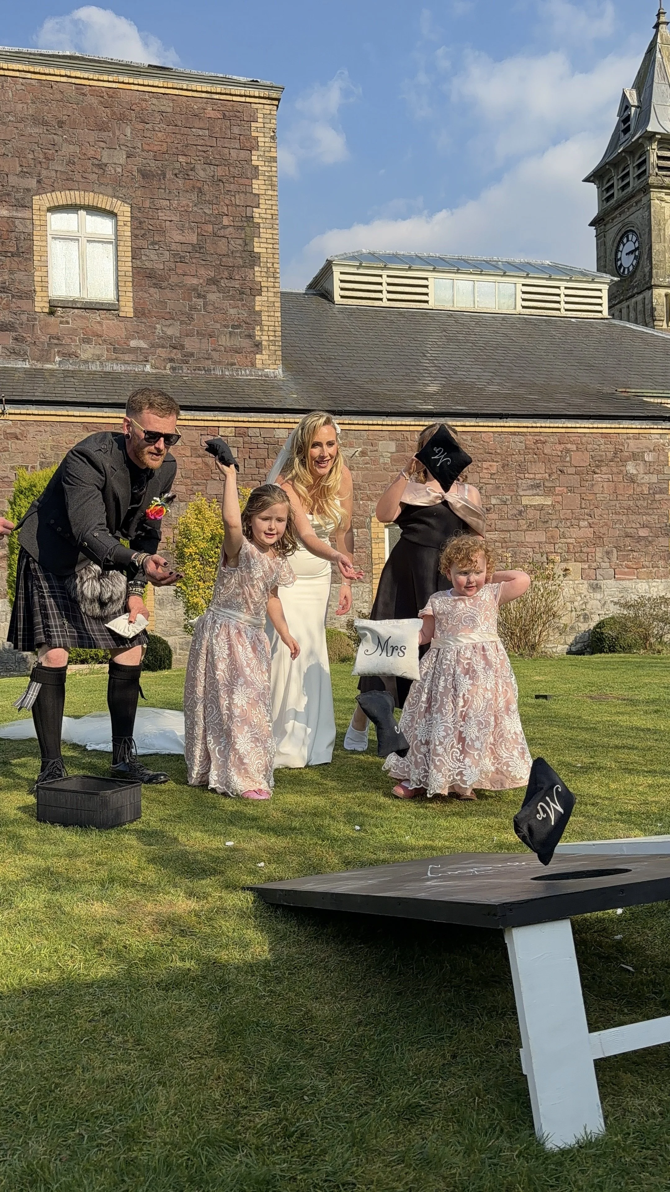 A wedding celebration outdoors with a bride, groom, two flower girls, and an officiant, all standing on grass near a brick building with a clock tower in the background, during sunny weather.