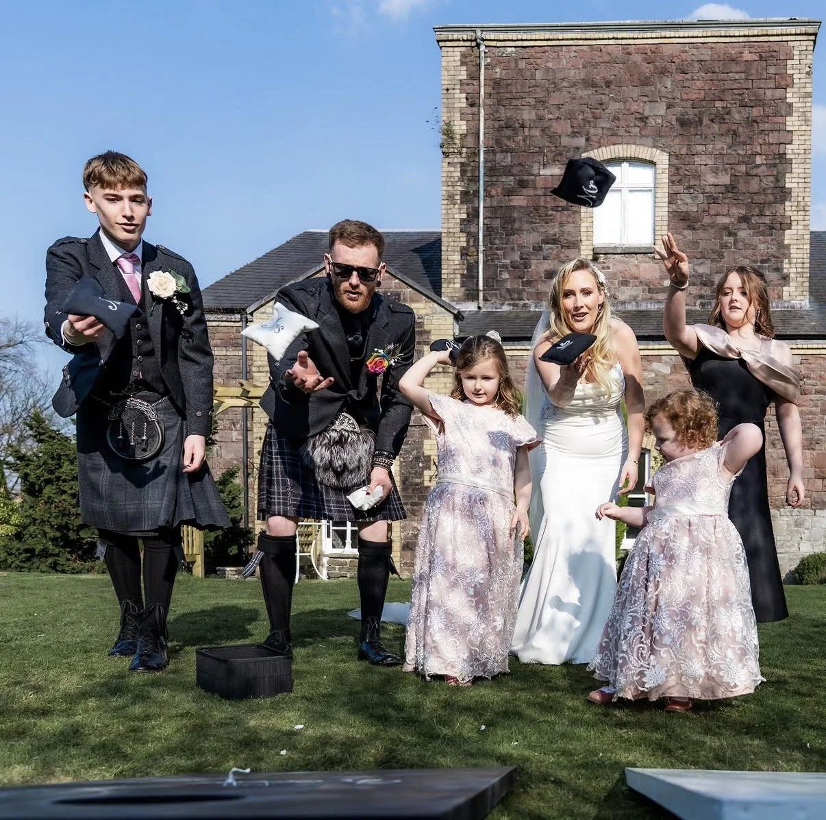 Group of people dressed in wedding attire outdoors, with some throwing confetti and others smiling, in front of a brick house.