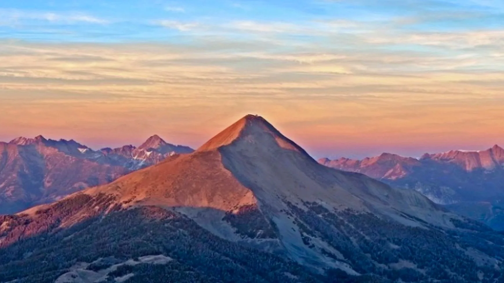 Sunset over mountain range with a prominent volcanic mountain in the foreground, surrounded by smaller peaks, under a sky with soft clouds.